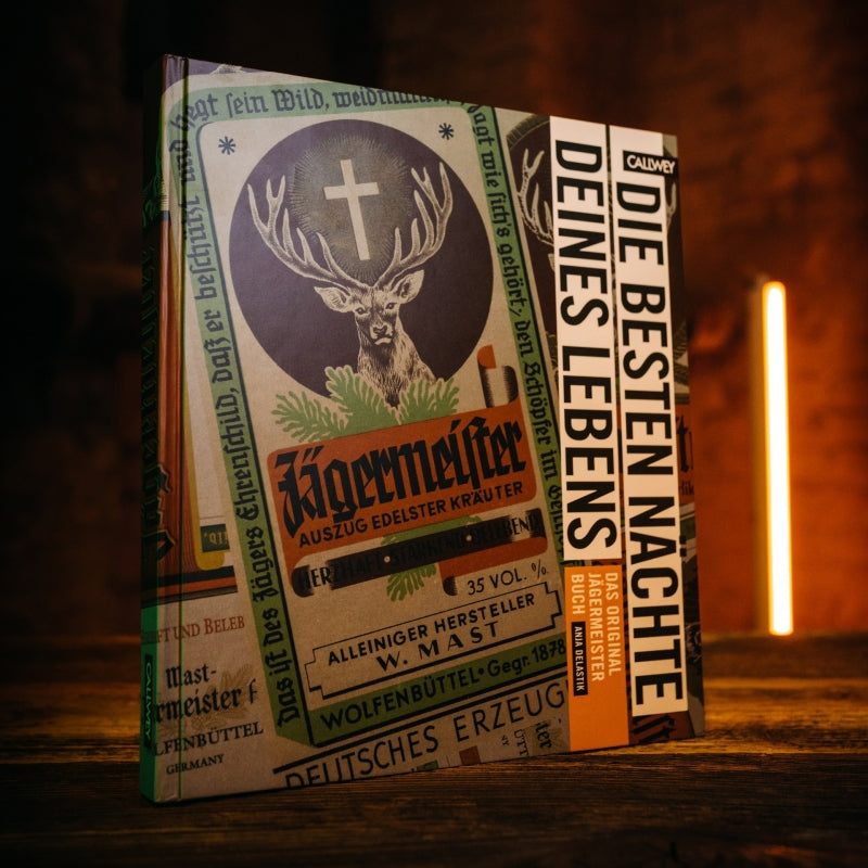 Book cover of Jagermeister Book, showing Jagermeister lables on top of each other, on a wooden table in a dark room with a single light