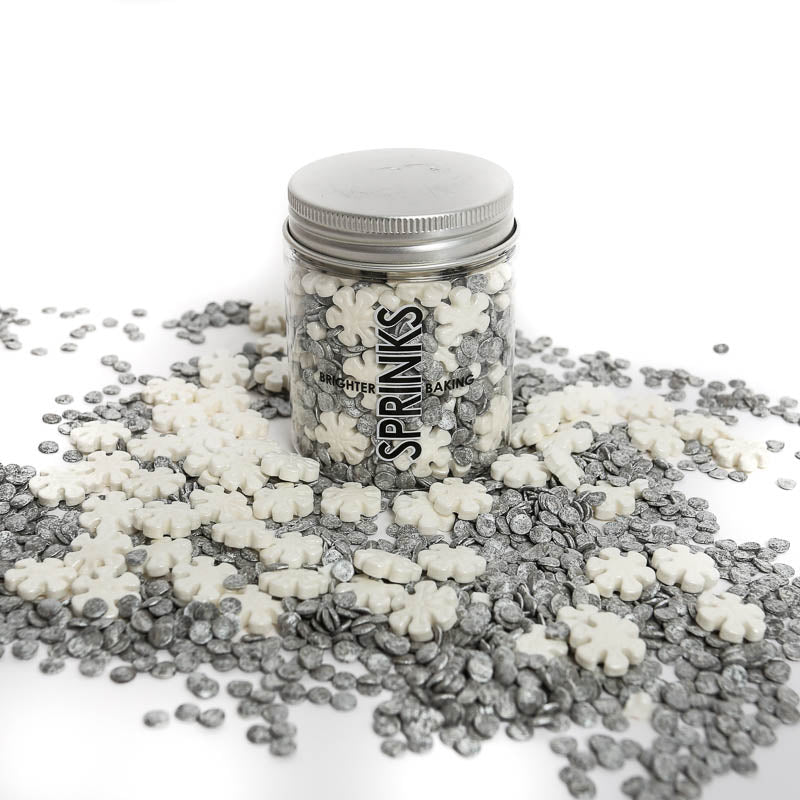 Image of container of white and silver snowflake shaped sprinkles for cake or cookie decorating shown with the snowflakes scattered around the container on a white counter