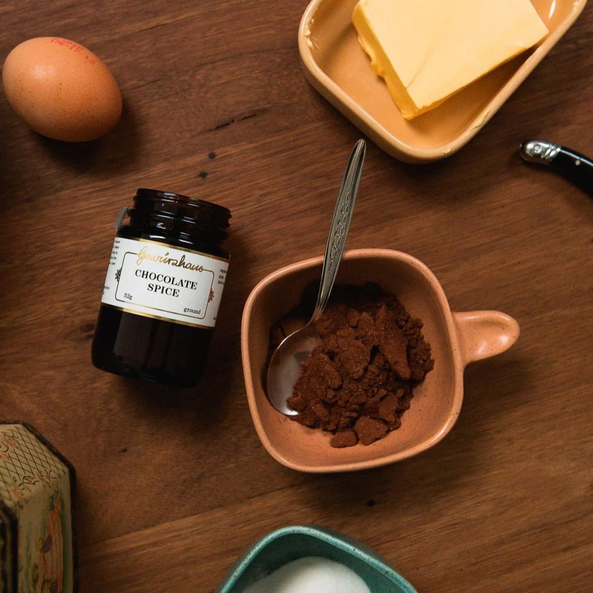 Photo of a jar of Gewürzhaus Chocolate Spice, displayed on a wooden table, with a spoon in an orange bowl filled with chocolate spice, an egg, a bowl of butter and a bowl of sugar.