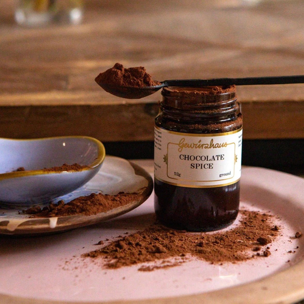 Photo of a jar of Gewürzhaus Chocolate Spice, displayed on a wooden table, with a heaped spoon of chocolate spice resting on the jar, on several stacked plates.