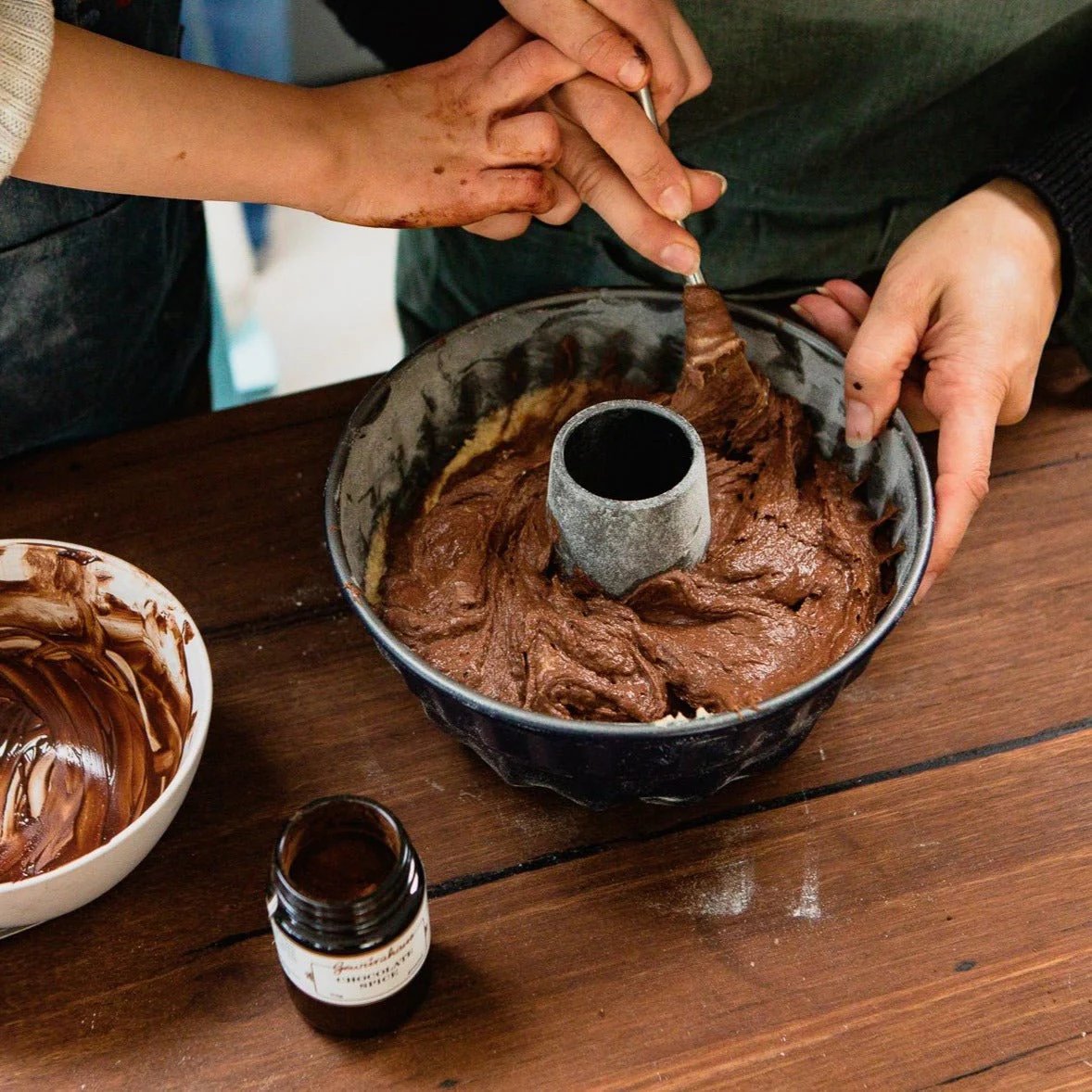 Photo of a jar of Gewürzhaus Chocolate Spice, displayed on a wooden table, with two people stiring cake batter in a bunt tin and a mixing bowl.