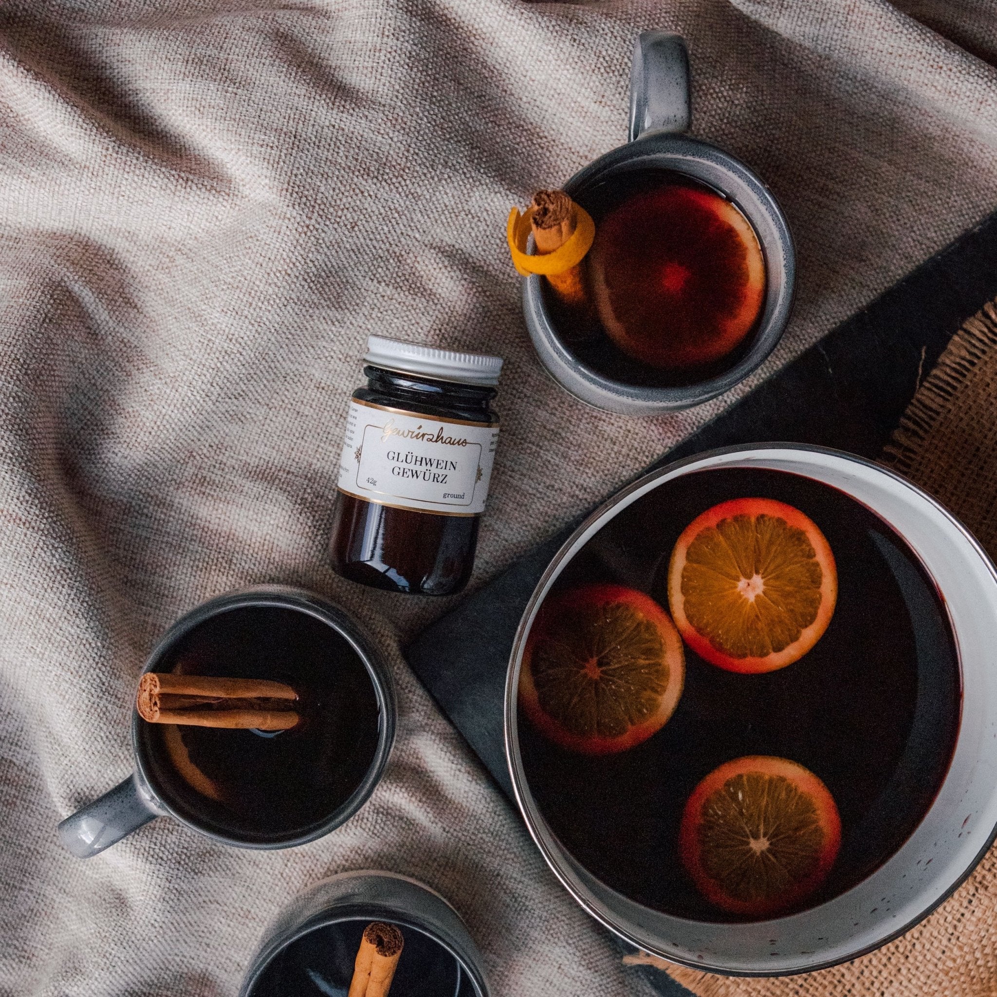 Photo of a jar of Gewürzhaus Glühwein & several mugs of Glühwein with citrus floating in the mugs. Shown against a beige linen tablecloth.