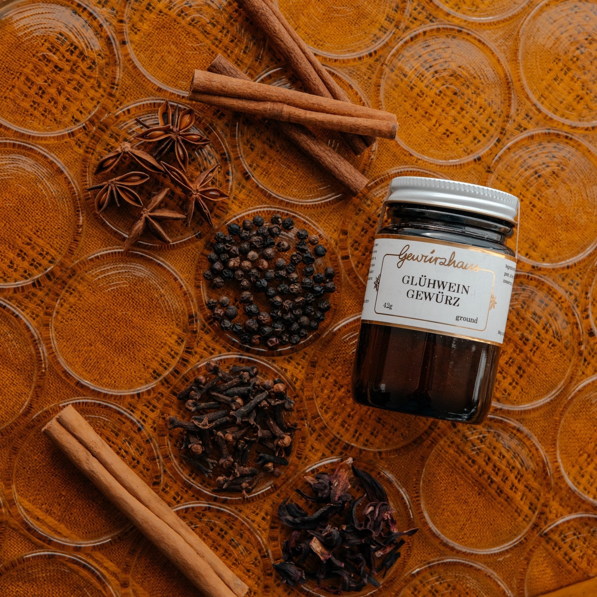 Photo of a jar of Gewürzhaus Glühwein, and 6 different dried spices. Shown against a orange tablecloth, with a clear glass laid over the top . 