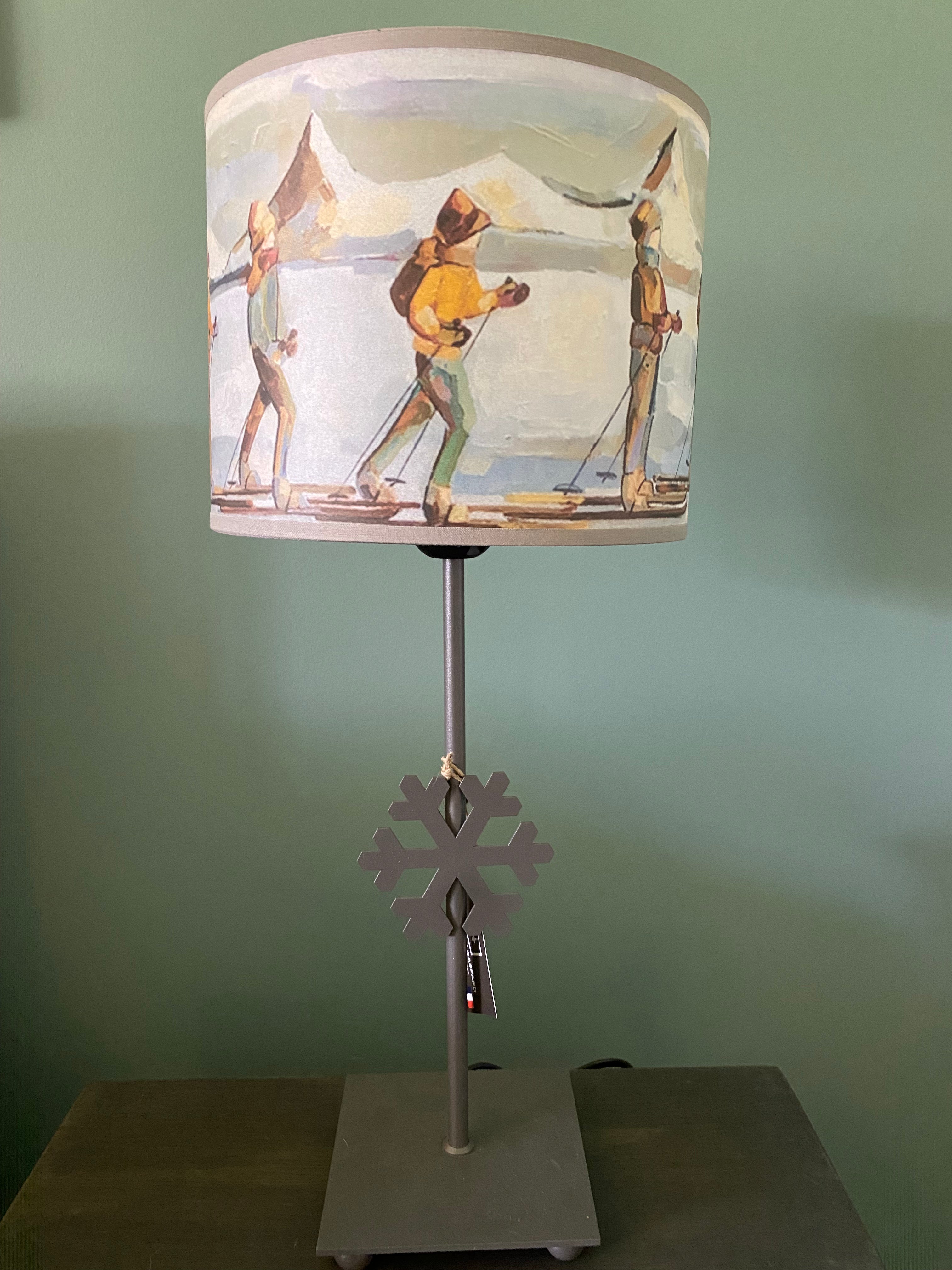 lamp with a square grey solid metal base supporting a rod with ca snowflake mid way up the rod, topped by a colourful canvas lamp shade. The shade depicts people on skis skinning across the snow in a line, one in front of the other, with snow capped mountains in the background. Sitting on a wooden table behind a green wall. Lamp shade not included.