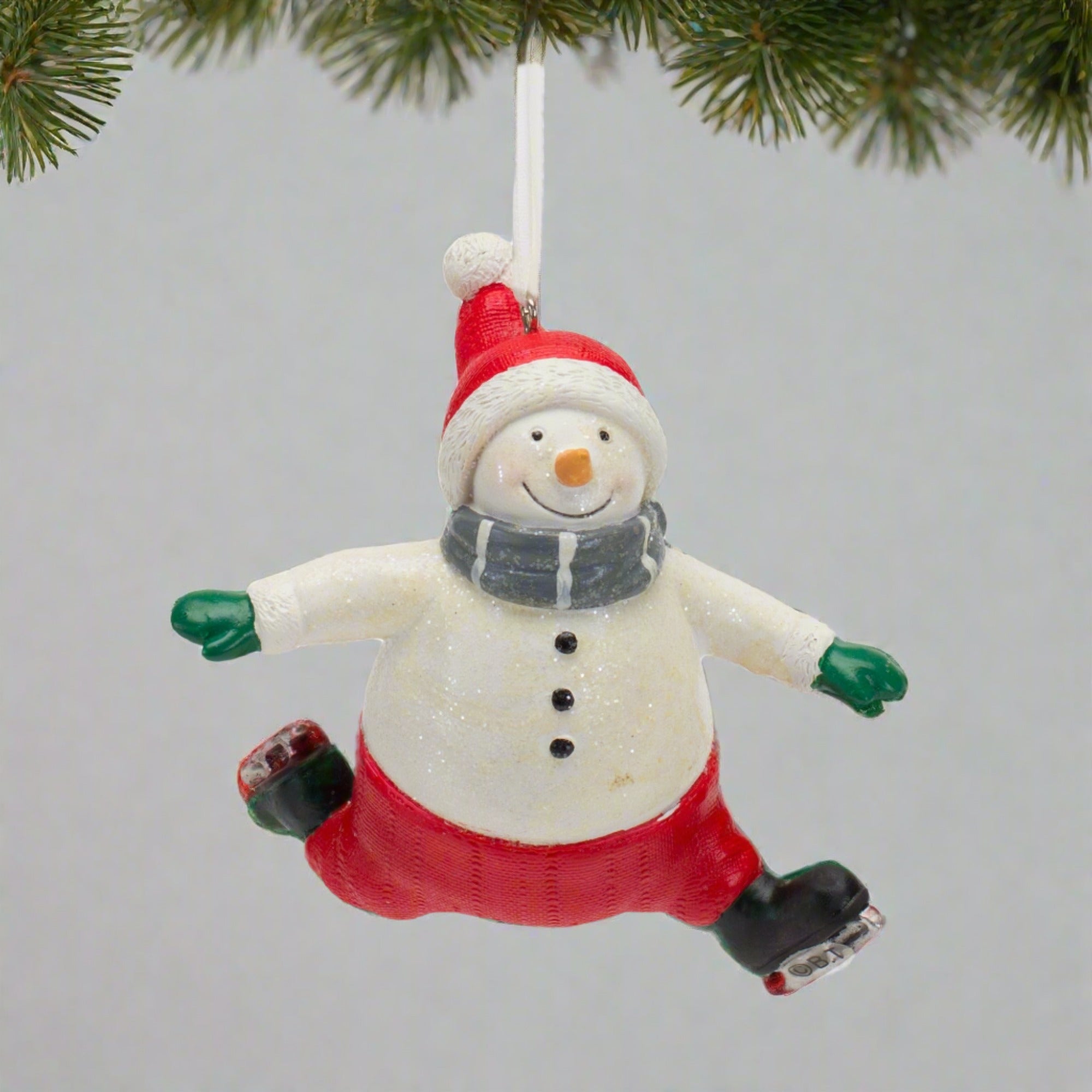 The snowman is wearing red pants, a red beanie with a white pom pom, green gloves, grey scarf and black skates. Hanging from a Christmas Tree