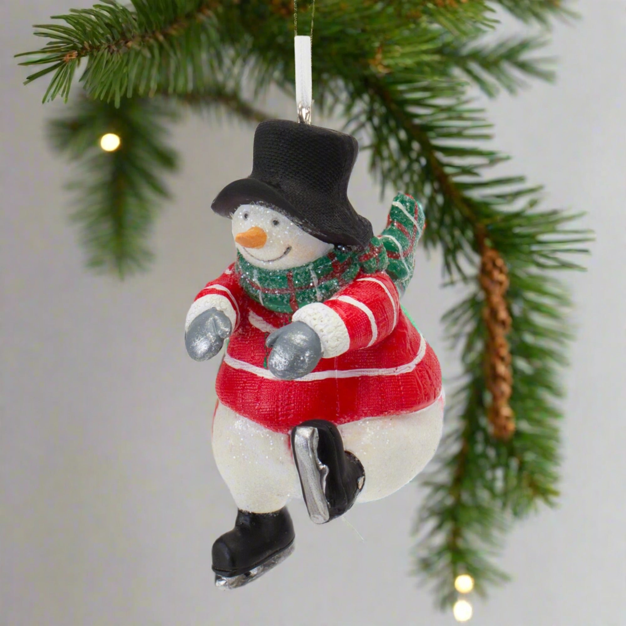 The Snowman is wearing a black top hat, red jumper with white stripes, green scarf, grey gloves and black skates. Hanging from a Christmas Tree