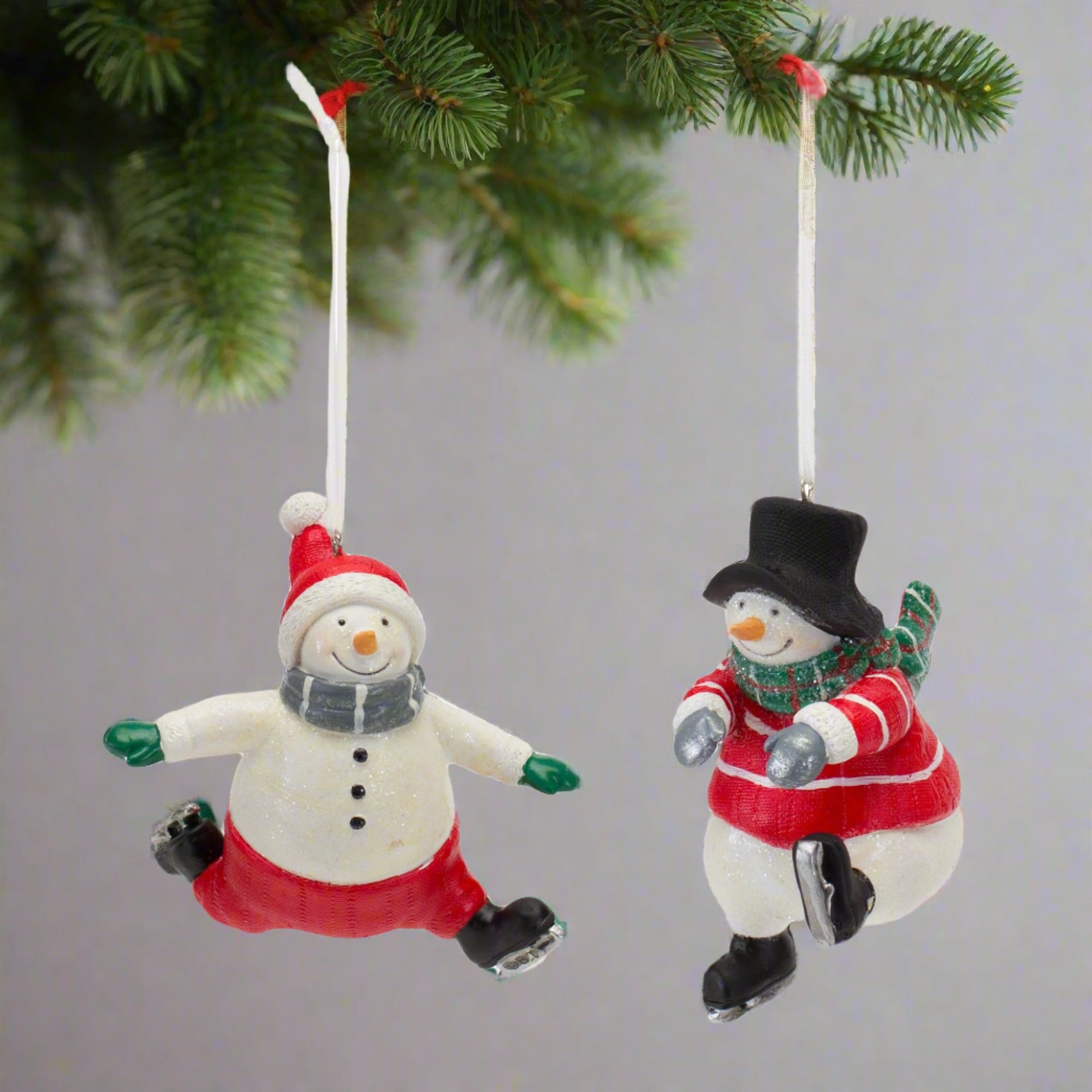 Skating snowman hanging ornaments. One is wearing red pants, a red beanie with a white pom pom, green gloves, grey scarf and black skates. The other is wearing a black top hat, red jumper with white stripes, green scarf, grey gloves and black skates. Hanging on a Christmas Tree