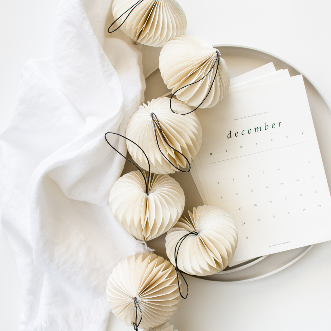 White Paper Christmas Ornaments in four different shapes: a heart, sphere and a jewel sitting on a white plate, next to a white December calendar page with a white serviette against a white background.