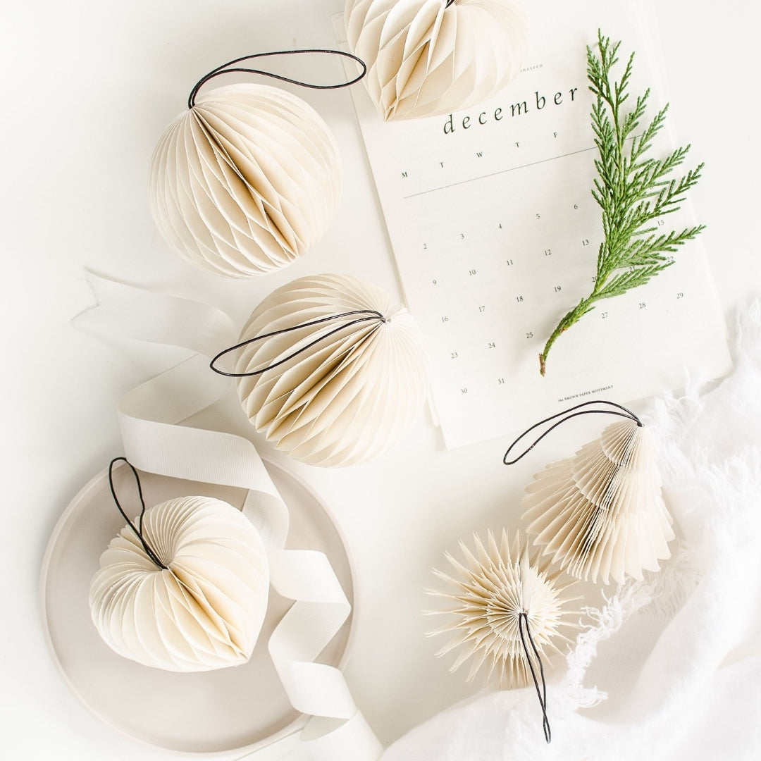 White Paper Christmas Ornaments in four different shapes: a tree, heart, sphere and a jewel sitting on a white plate, next to a white December calendar page with a white serviette and ribbon, & a sprig of pine against a white background.