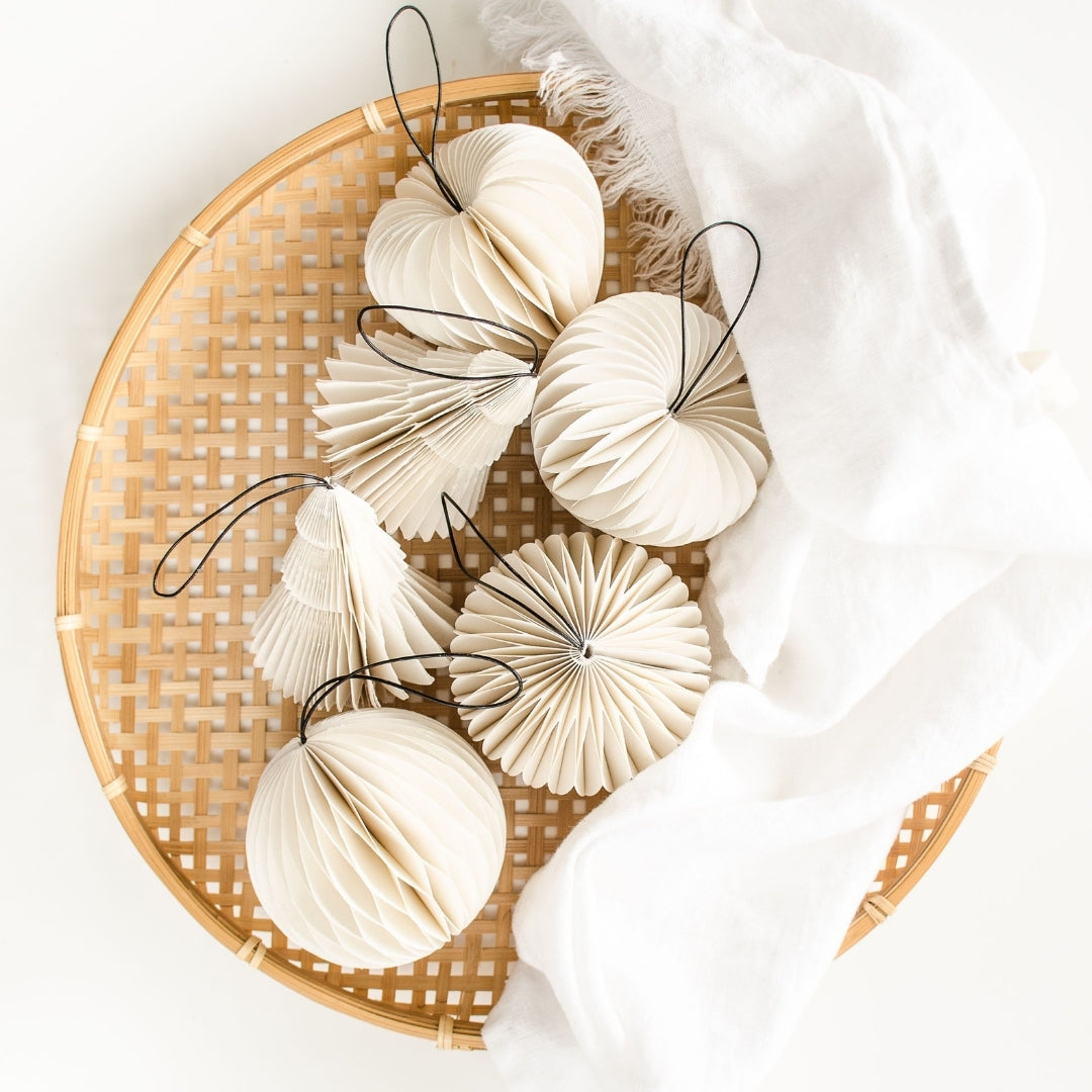 White Paper Christmas Ornaments in four different shapes: a tree, heart, sphere and a jewel sitting in a wicker basket with a white serviette against a white background.