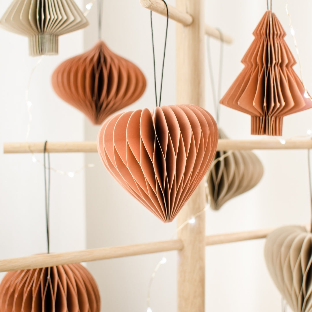 Clay and Linen Coloured Paper Christmas Ornaments in the shapes of trees, hearts and jewels hanging on a wooden frame against a white background. 
