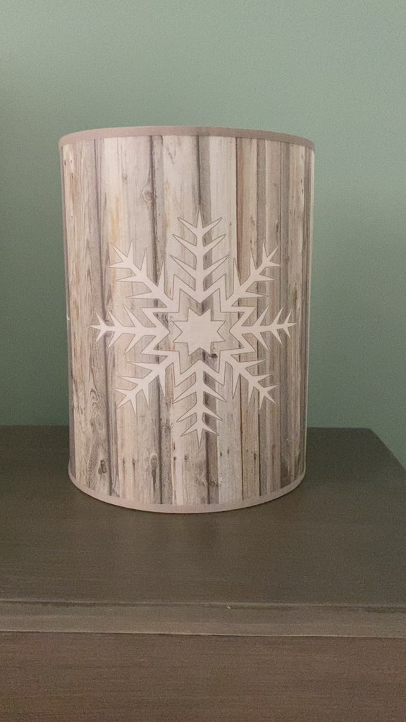 Video showing A cylindrical table lamp made of a cylinder of canvas printed with a photo of wood grain, with a white snowflake embossed on opposing side of the lamp, against a green painted wall on a wooden table. Lamp is being rotated from side to side to show it at different angles.