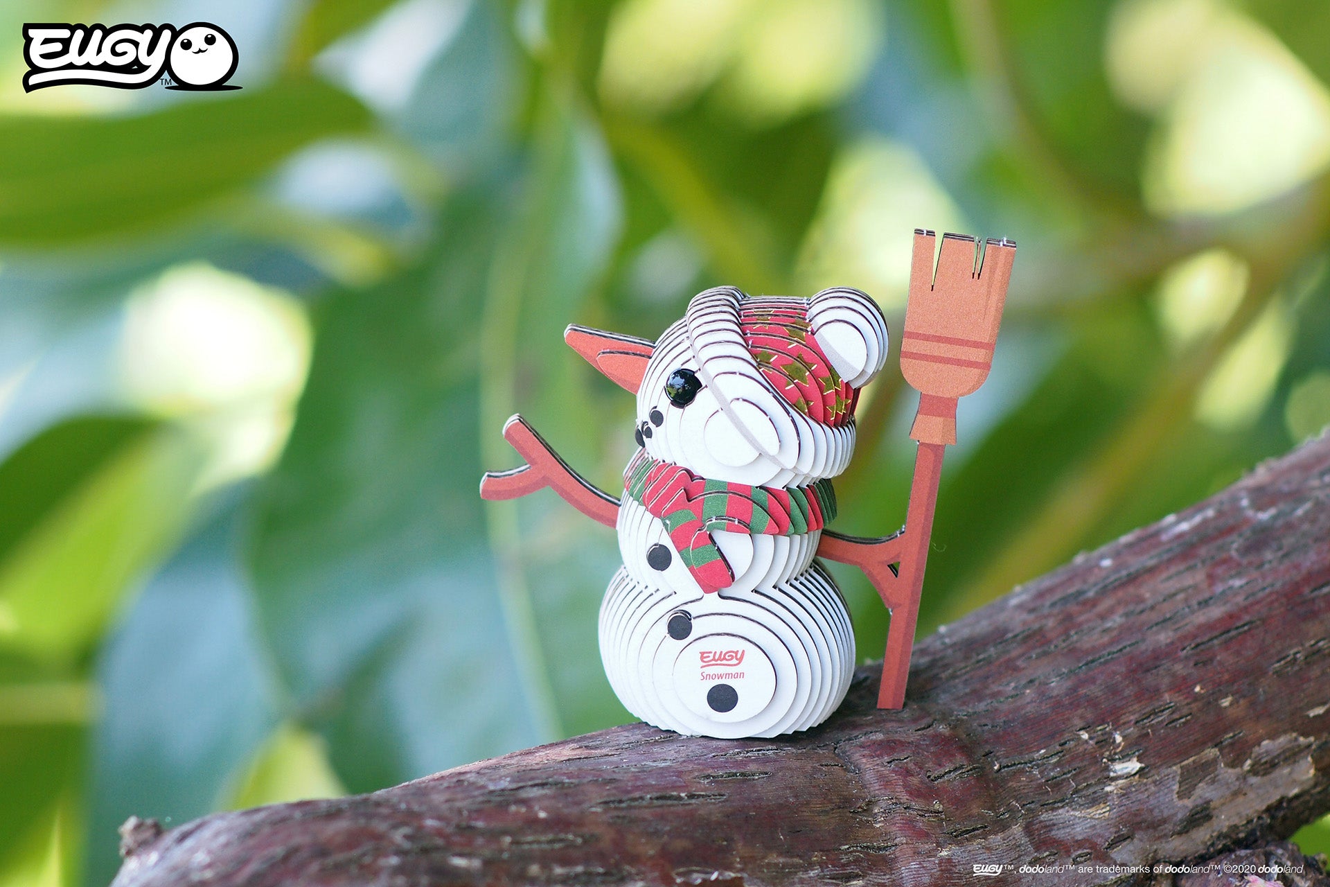 Image of an EUGY Snowman, facing left but viewed from the front & sitting on tree branch in front of a blurred leafy background