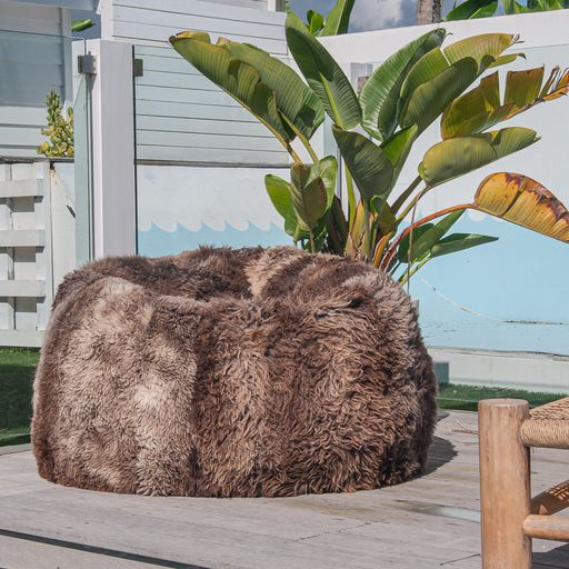 Beautiful brown Long Wool Sheepskin Bean Bag sitting outside on a wooden deck and in front of a glass pool fence and a large potted bird of paradise plant