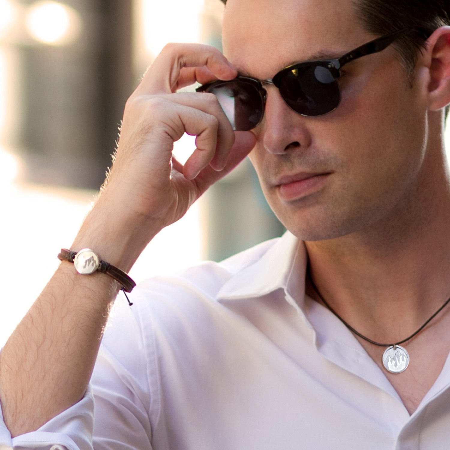 Image shows a close up of a man with brown hair, wearing a white shirt and sunglasses. He is adjusting his sungalsses and showing a stainless steel round pendant with mountains engraved into their surface with leather thongs attaching the pendant as a bracelet, and the same pendant as a necklace hanging by a leather thong.
