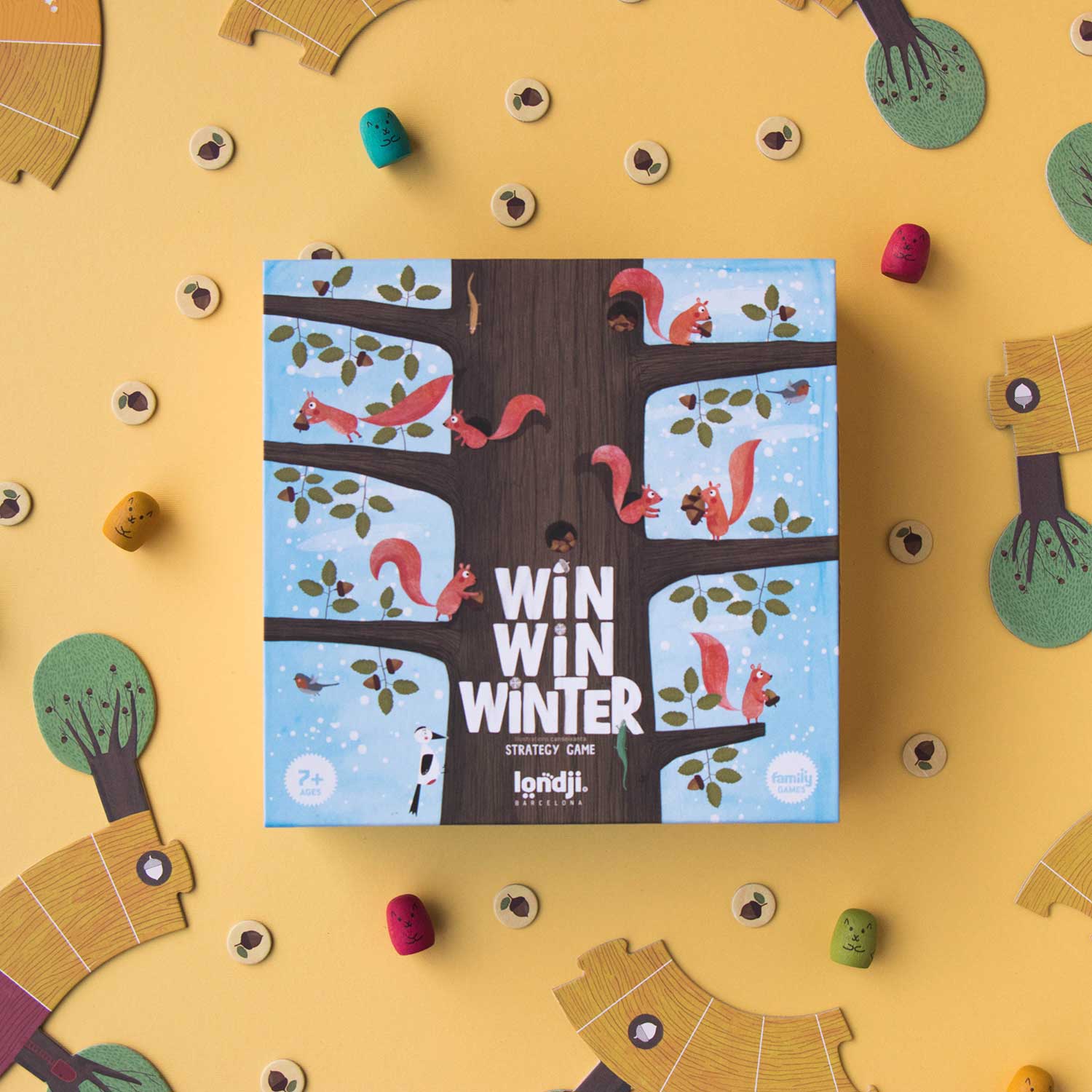 Image shows an overhead view of the Win Win Winter game box, illustrated with a tall tree with many squirrels scampering across the branches. The box is surrounded by board game pieces. Shown against a yellow backdrop.