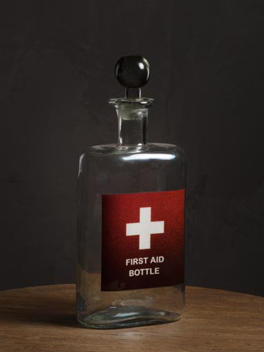 Glass Decanter with the red and white First Aid symbol labels on its front & with glass stopper, on a wood table and grey wall