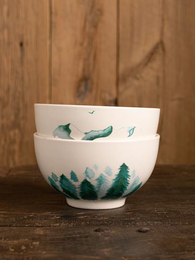 Pair of white porcelain bowls painted with trees and mountains in green, stacked on top of each other and sitting against a wood panelling wall and floor