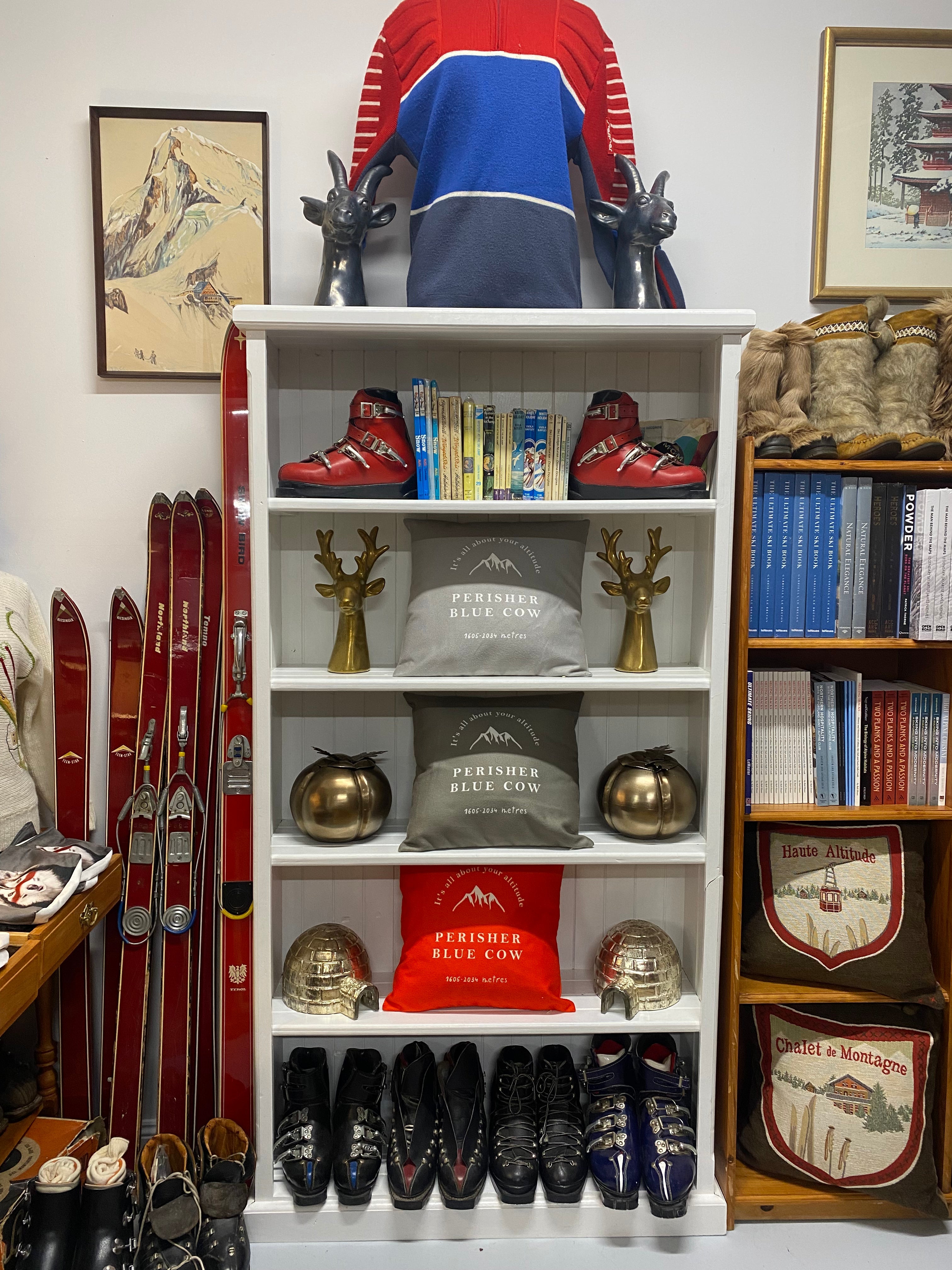 Image of white bookshelf, surrounded by red vintage skis, many nooks and leather ski boots and with red, grey and taupe cushions printed with the words Perisher Blue Cow visible on the cushion and a drawing of a mountain