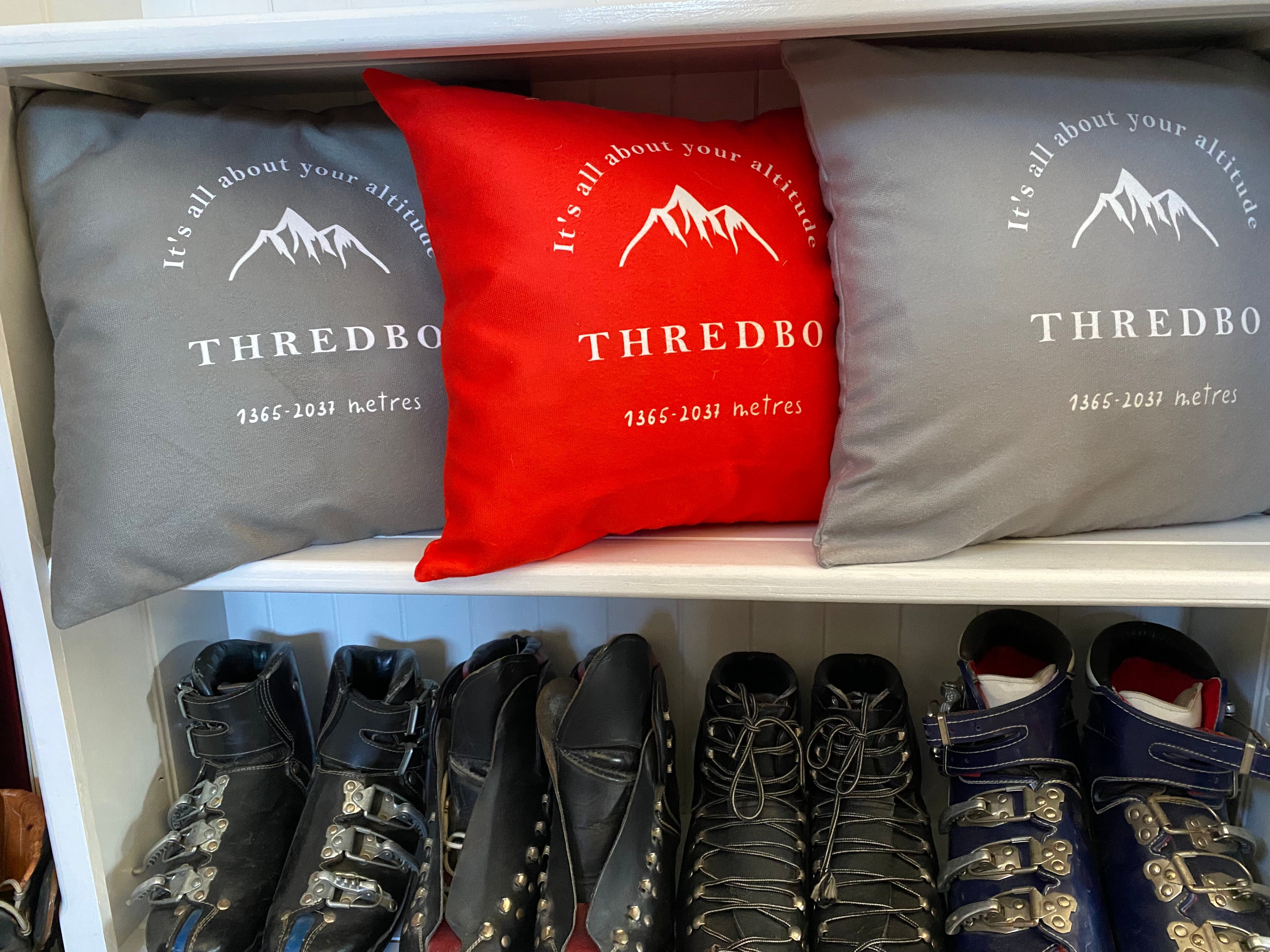 Image of white bookshelf with red, grey and taupe cushions printed with the words Thredbo; it's all about your altitude visible on the cushion and a drawing of a mountain, and vintage leather ski boots underneath
