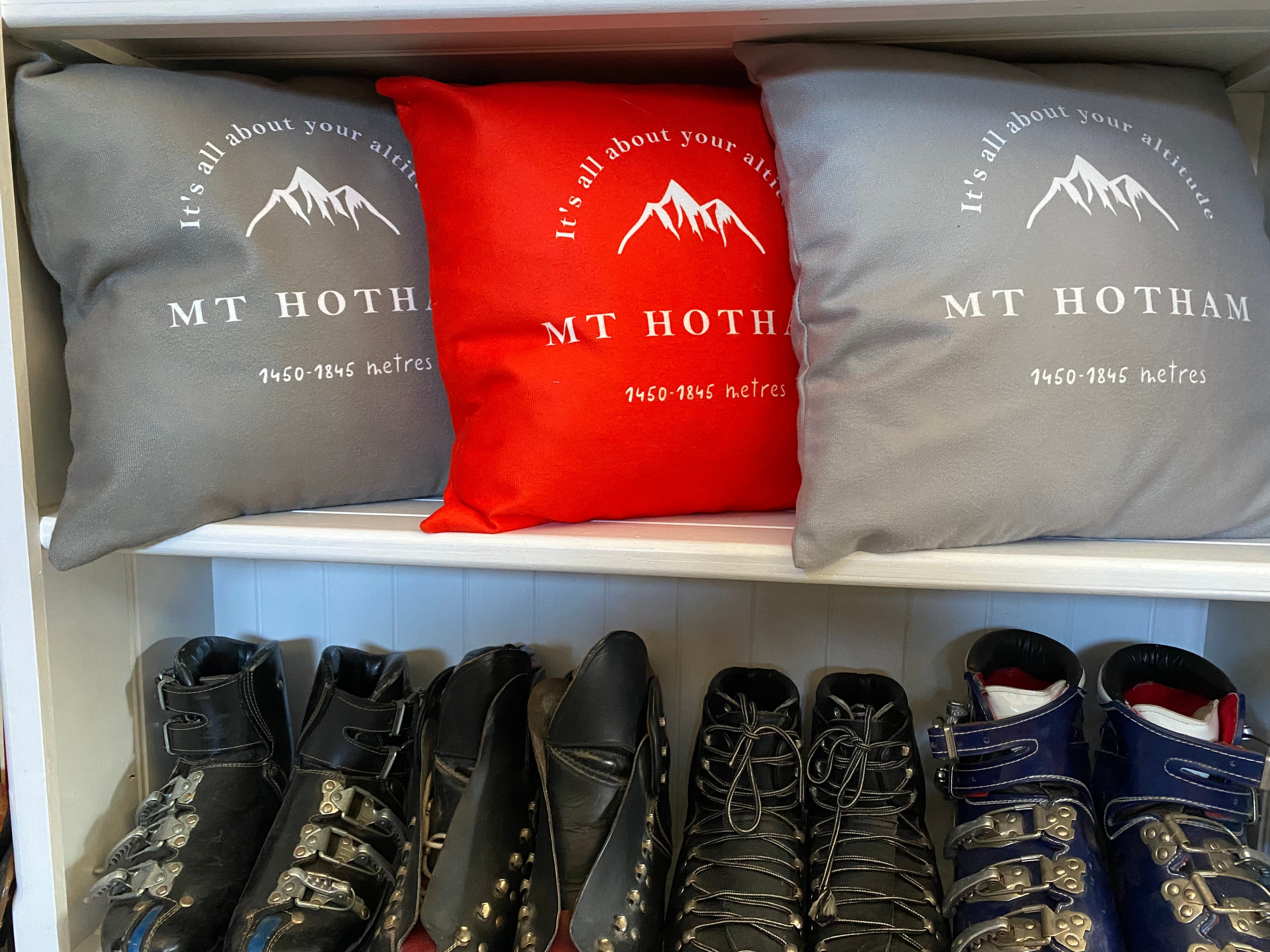 Image of white bookshelf with red, grey and taupe cushions printed with the words Mt Hotham; it's all about your altitude visible on the cushion and a drawing of a mountain, and vintage leather ski boots underneath