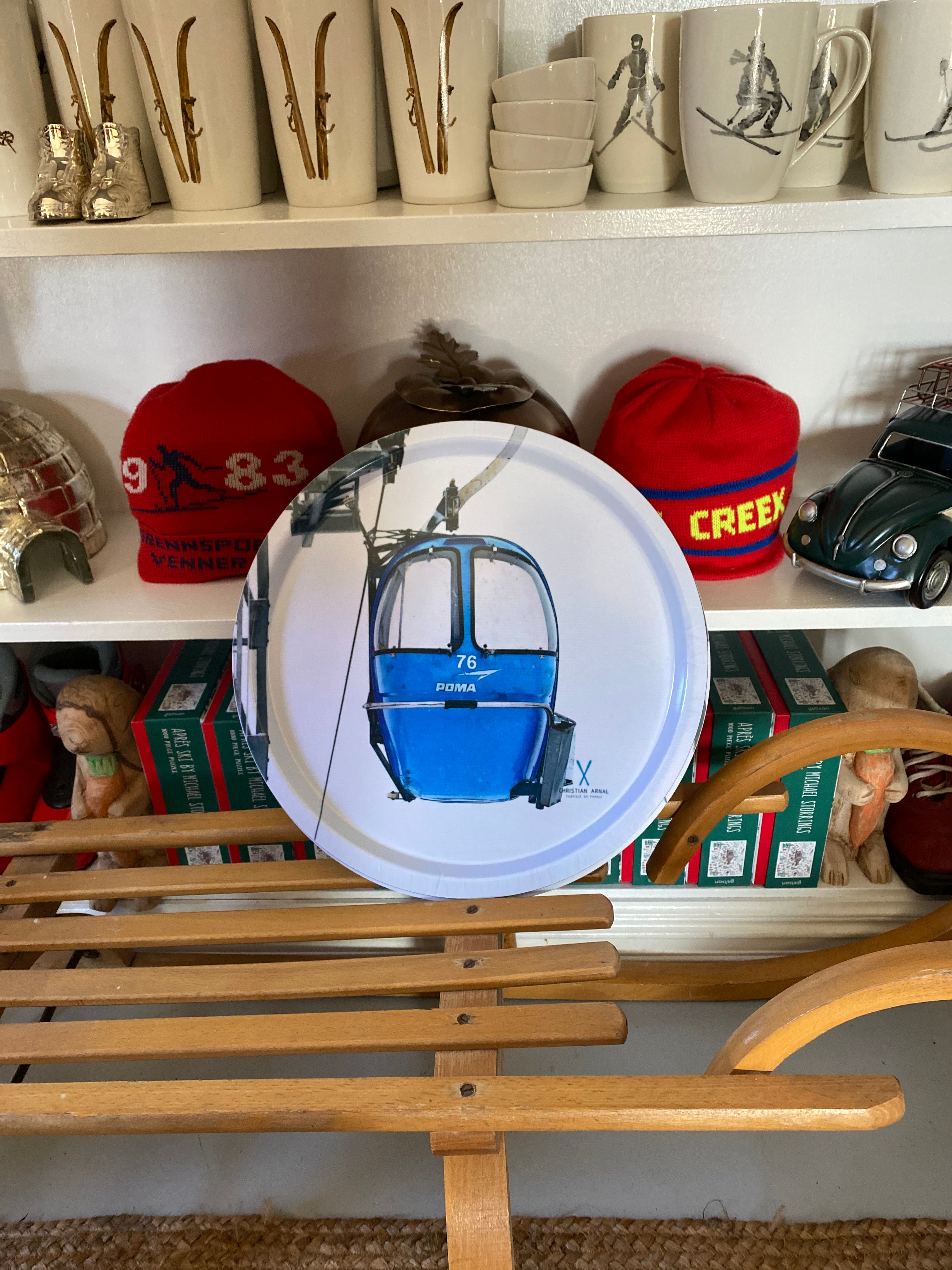 Photo of a Blue Vintage Gondola printed onto a Round White Serving Tray, resting on a vintage wooden sled in front of a white bookshelf filled with snow related products
