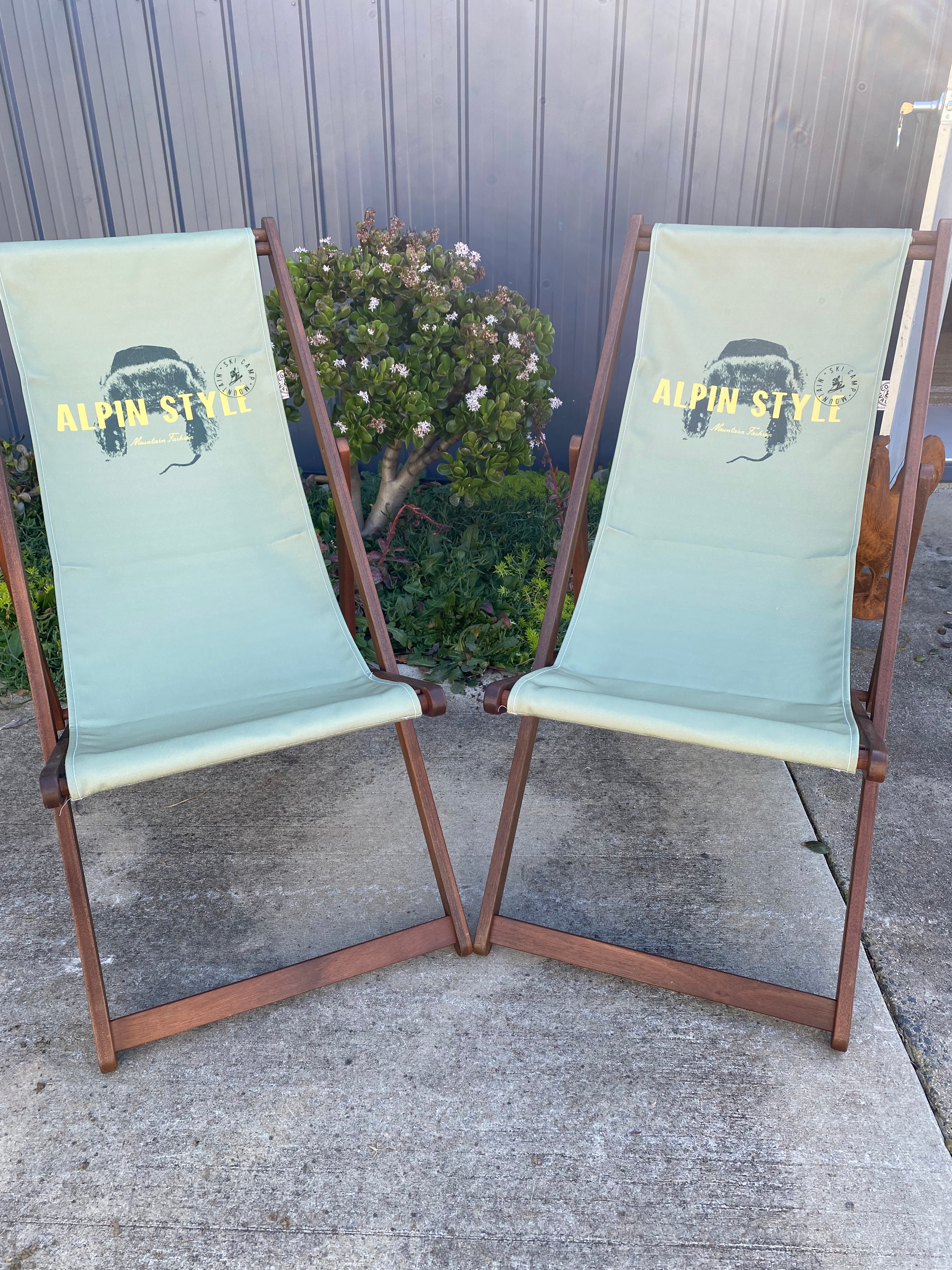 Pair of Coast & Valley Deck Chairs: Alpin Style design showing words in yellow with fur hat on a plain eucalyptus green background. 2 chairs facing forward. On a concrete floor against a blue steel shed and door with garden bed in the background.