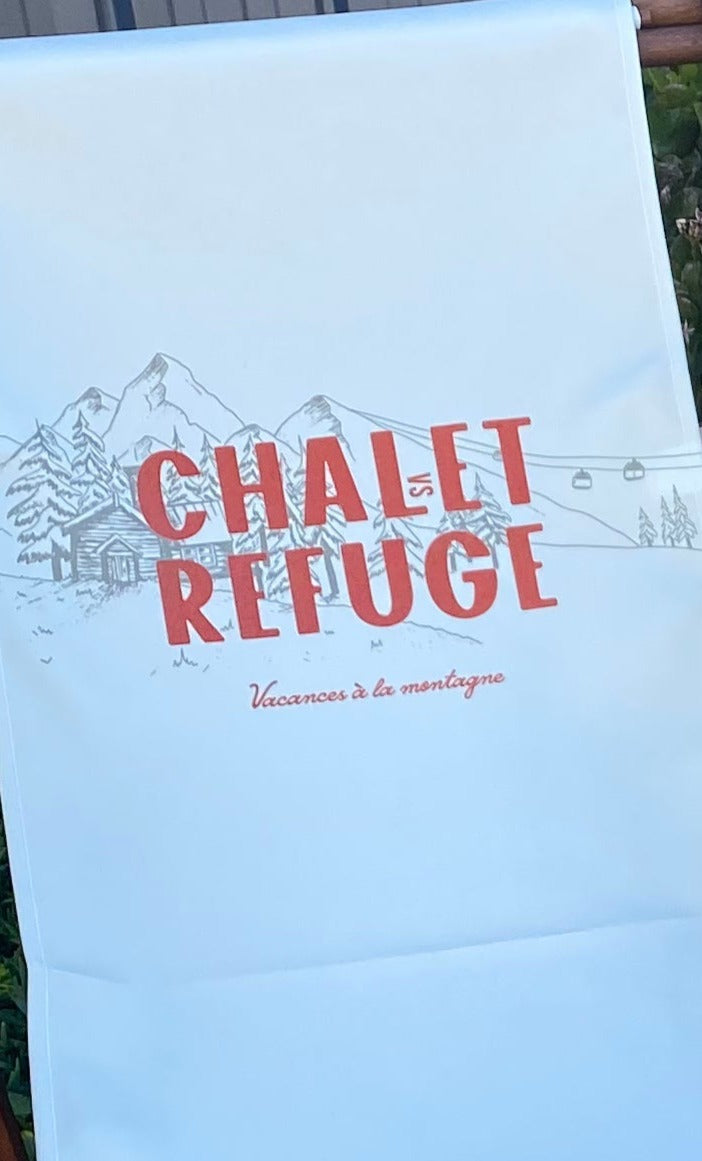 Front View of 2 Wooden deck chair frame with off-white canvas sling printed with an image ofa snowy mountain and a chalet building and the words Chalet vs Refuge written in red. Chairs sitting on concrete in front of a plant filled garden bed & blue corrugated iron wall.