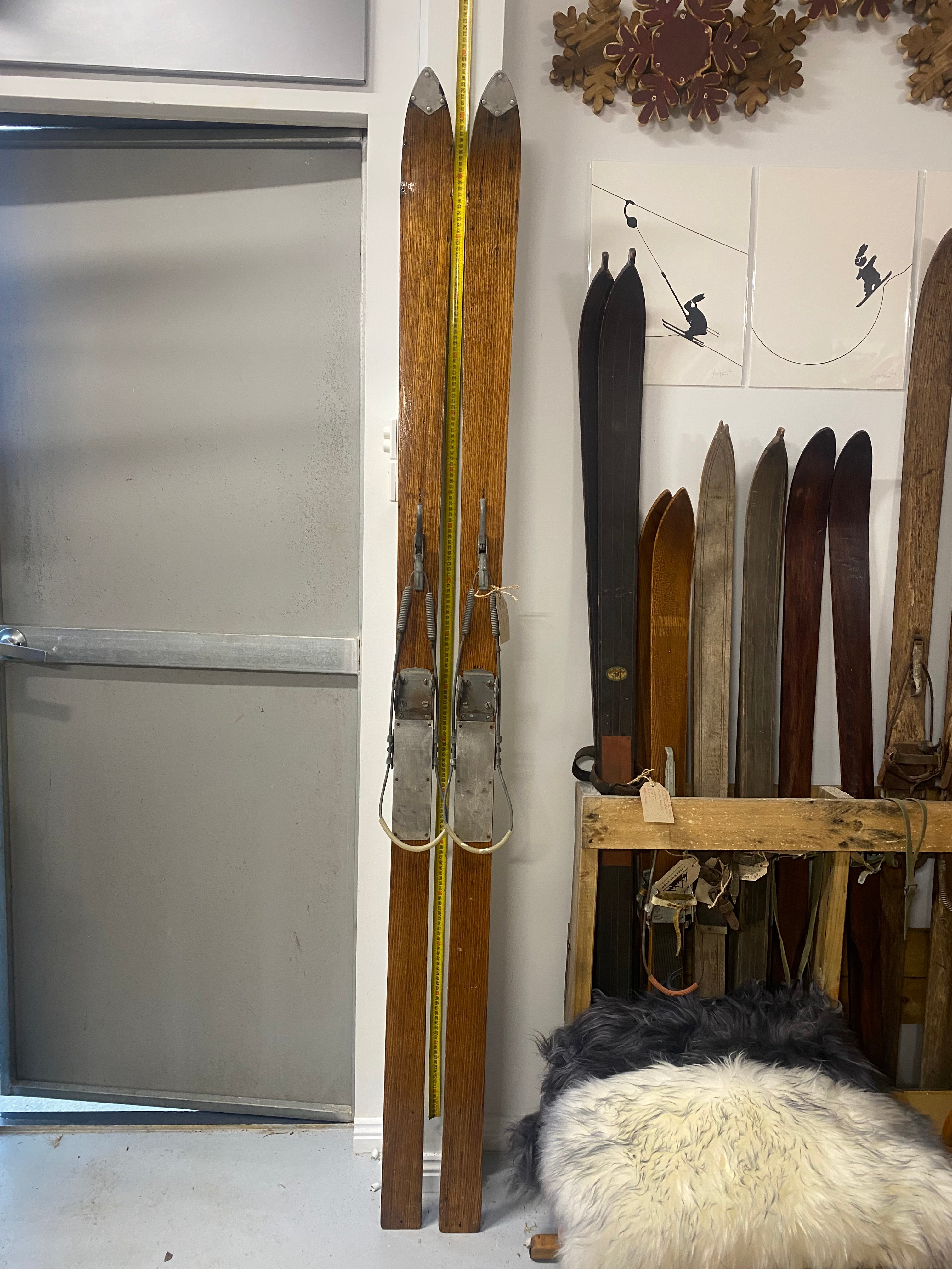Full Height Front View: Vintage Wooden Skis with metal bindings. Lent against a white wall, with more vintage wooden skis stacked to the right hand side, sheep skins on the floor and posters above the skis.