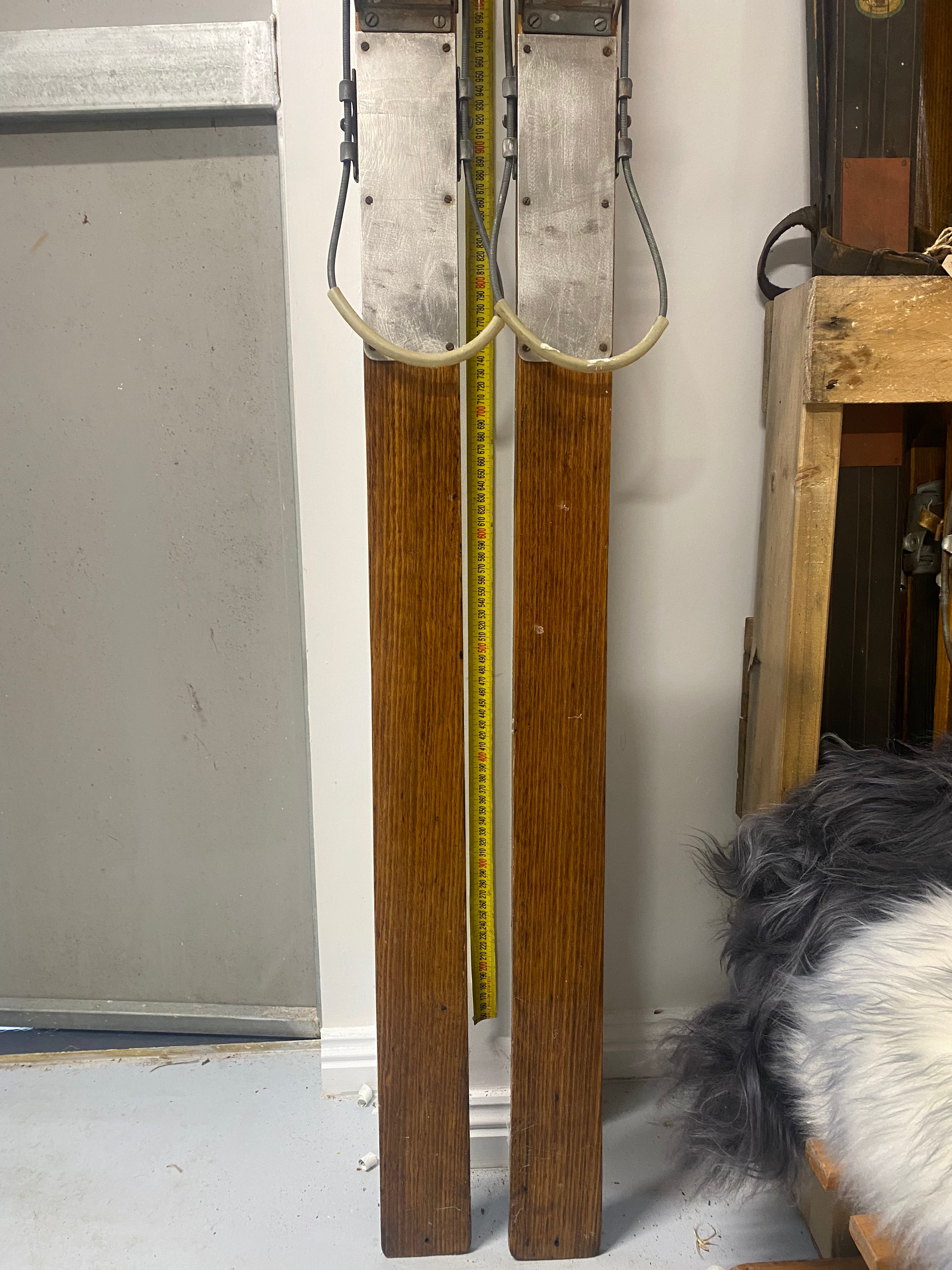 Bottom Half of Skis Front View: Vintage Wooden Skis with metal bindings. Lent against a white wall, with more vintage wooden skis stacked to the right hand side, sheep skins on the floor.