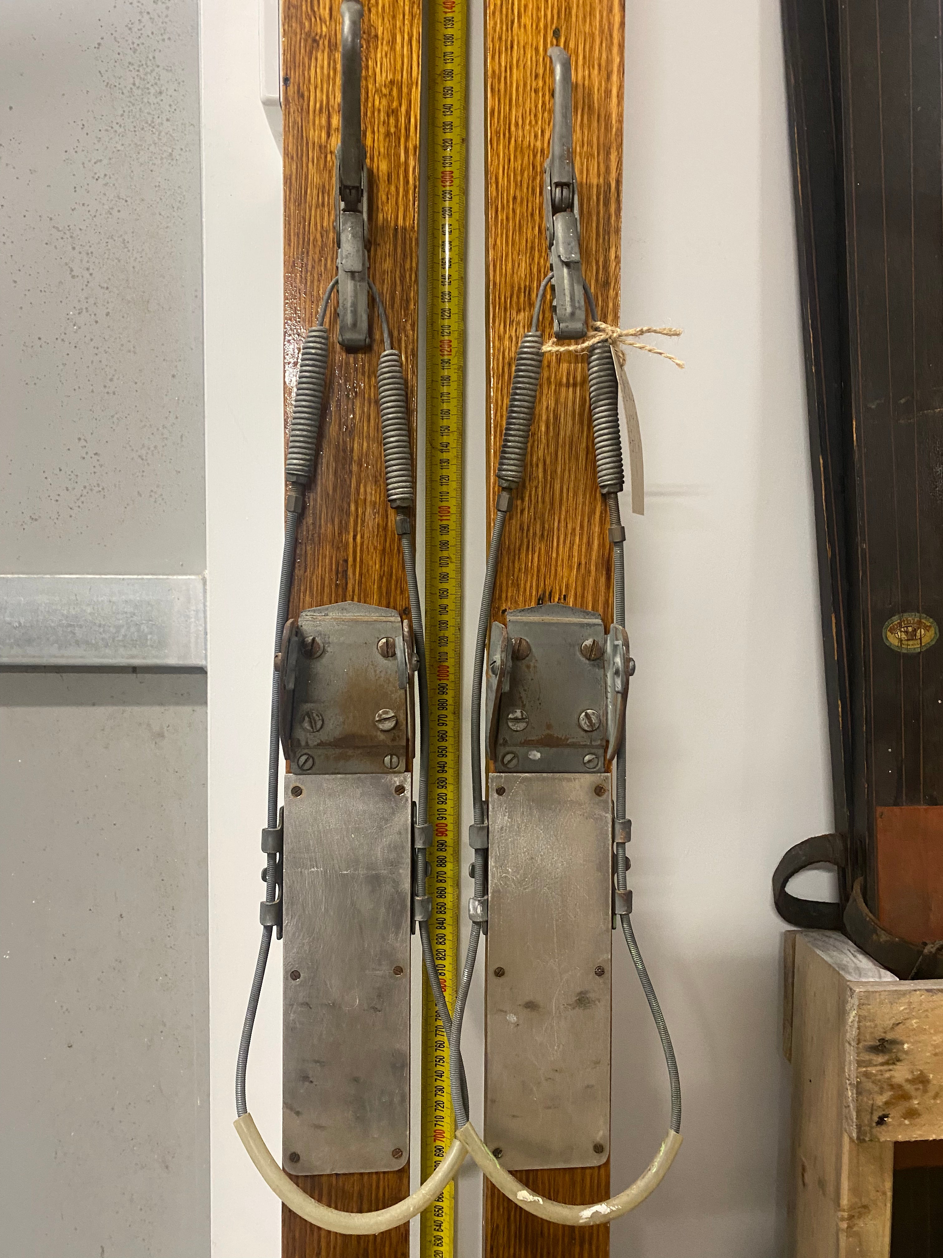 Ski Bindings Front View: Vintage Wooden Skis with metal bindings. Lent against a white wall, with a tape measure behind the skis