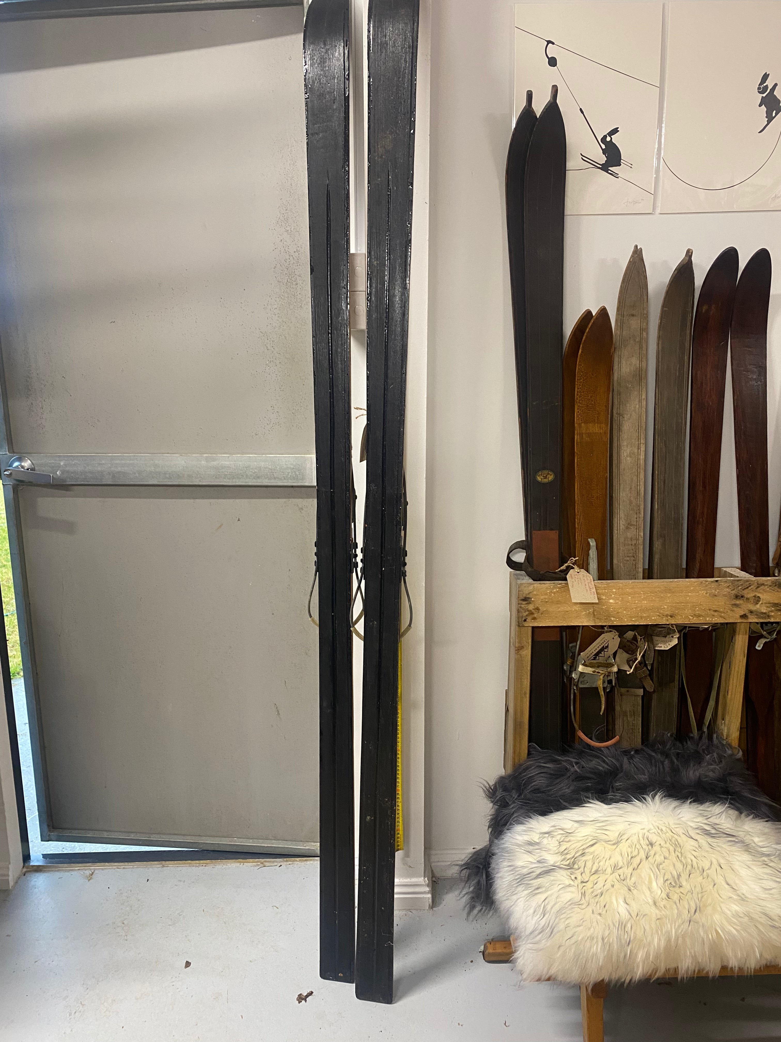 Bases View Full Height: Vintage Wooden Skis with metal bindings. Lent against a white wall, with more vintage wooden skis stacked to the right hand side, sheep skins on the floor and posters above the skis.