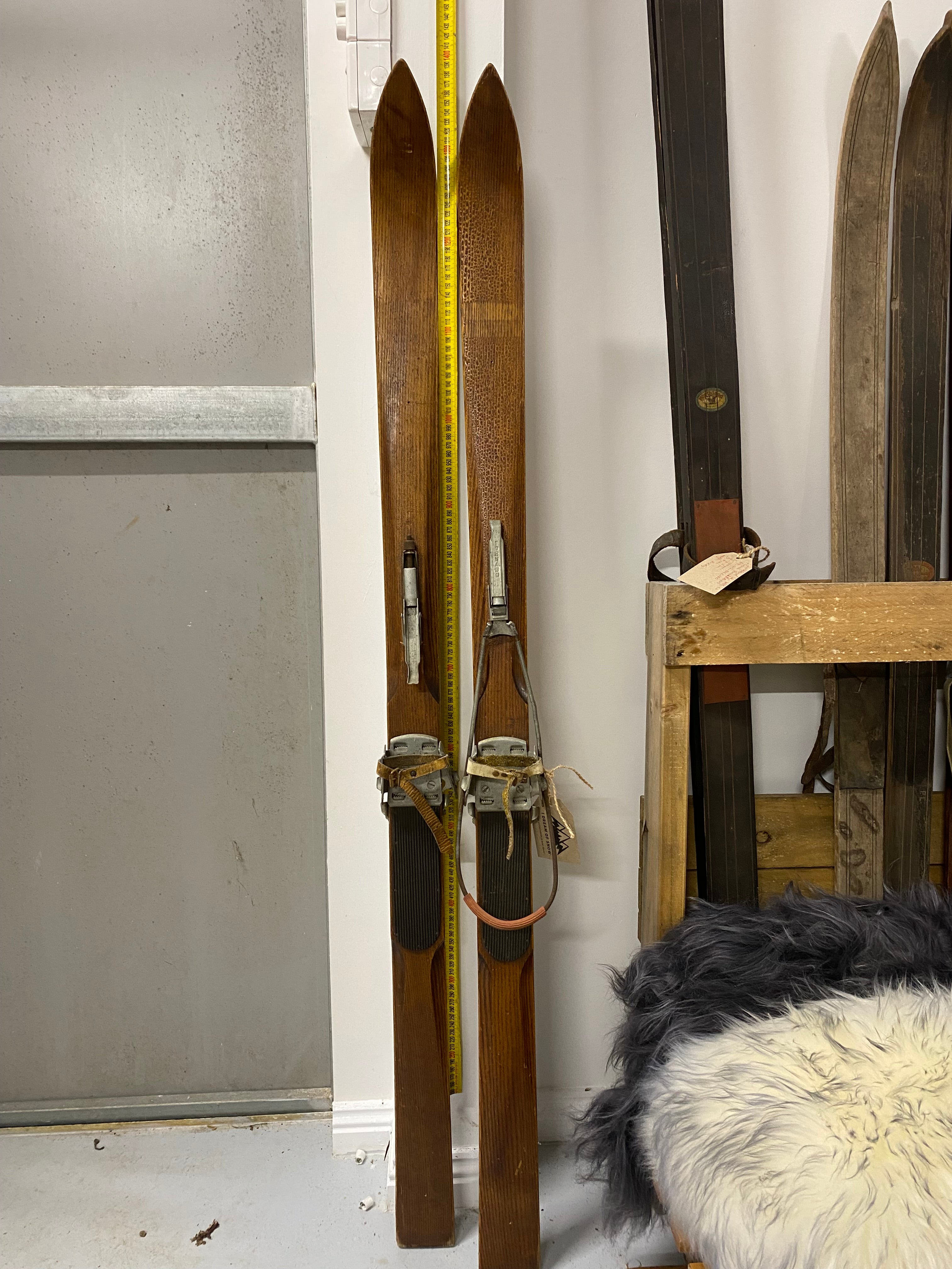 Full Height front view: unbranded vintage wooden skis with Tornado metal bindings. leaning against white painted wall with yellow measuring tape. On the right hand side are a number of stacked vintage skis.