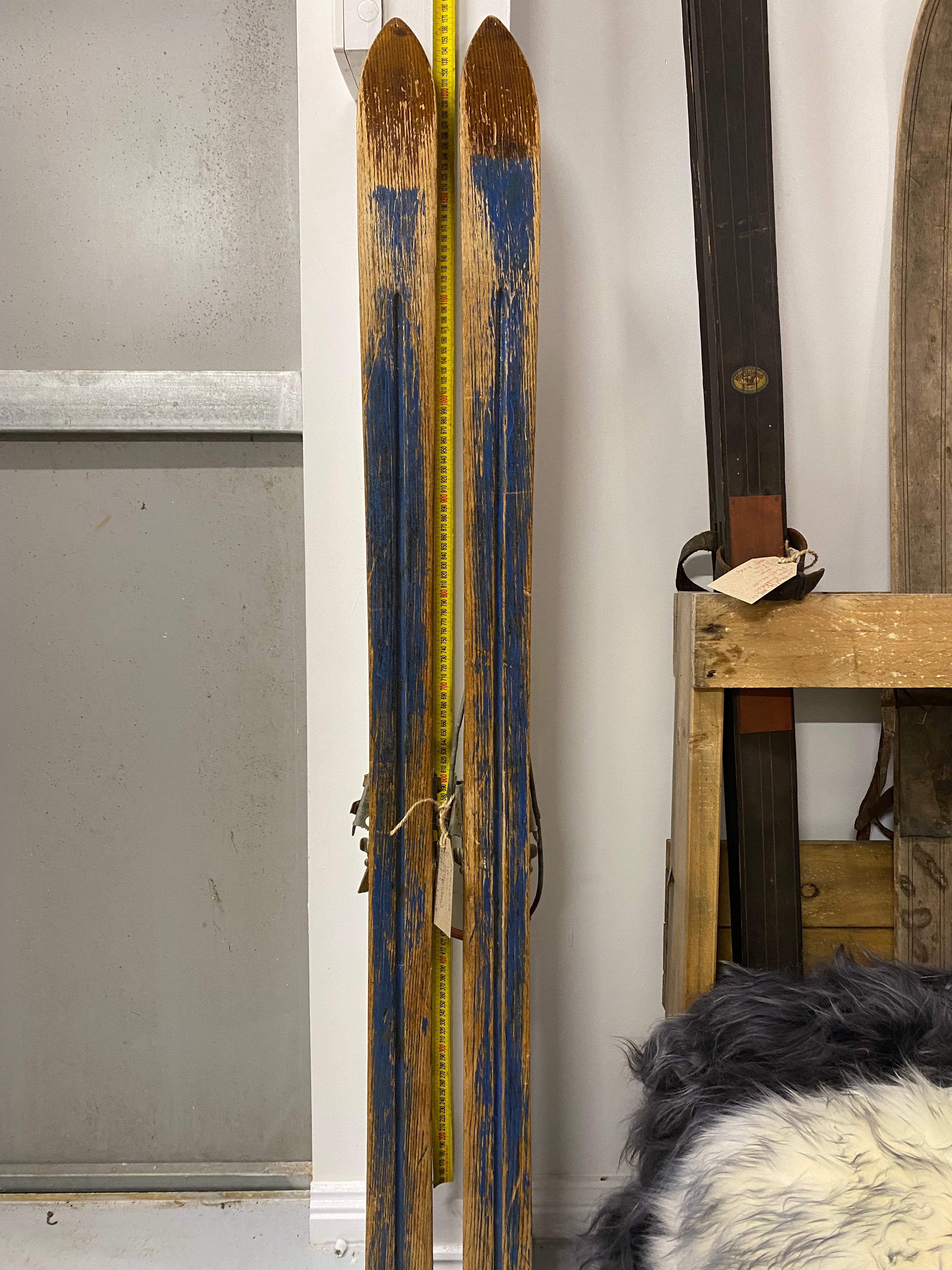 Full Height bases view: unbranded vintage wooden skis with partially painted blue bases. leaning against white painted wall with yellow measuring tape. On the right hand side are a number of stacked vintage skis.