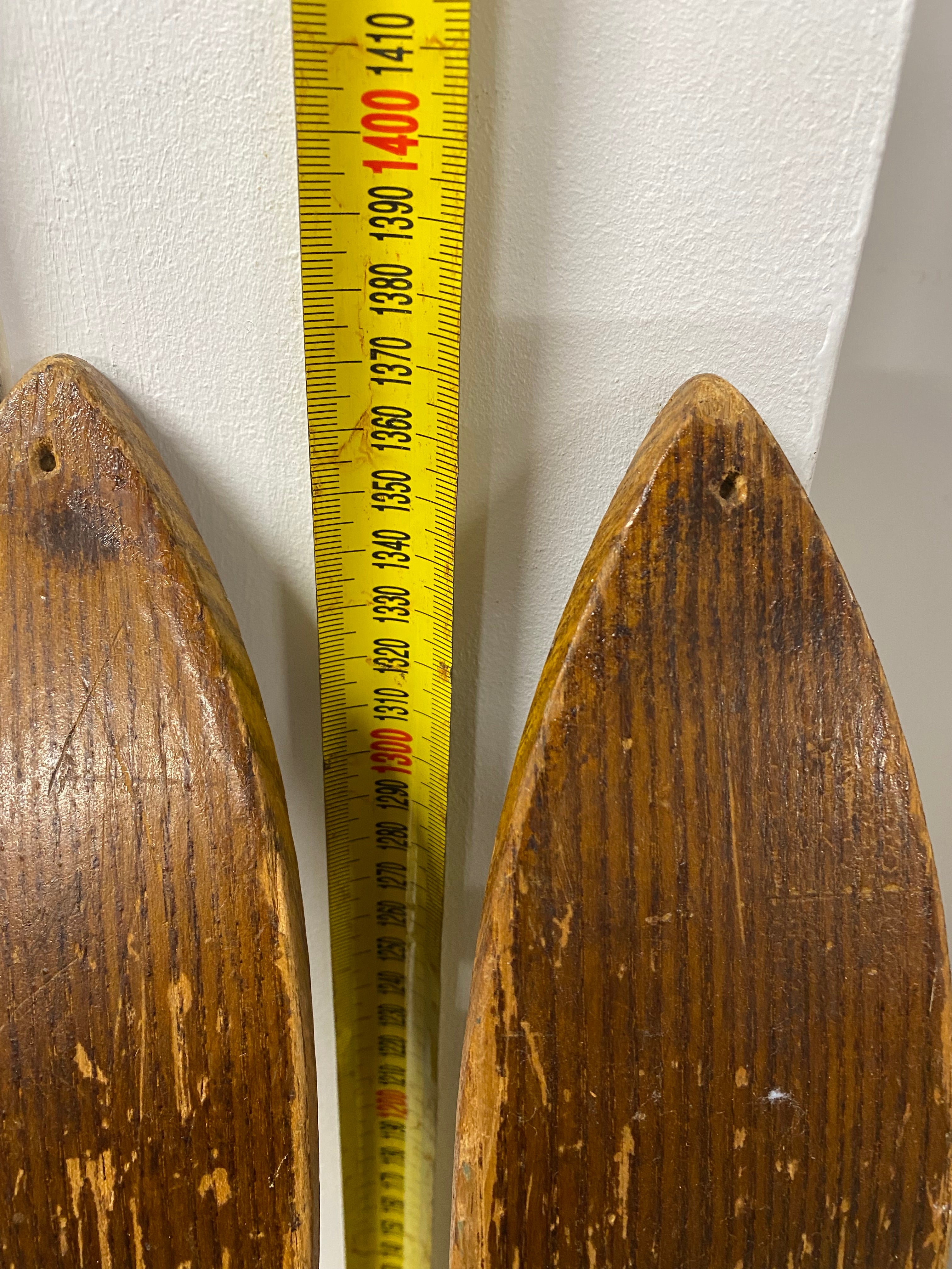 View of ski tip bases. Unbranded vintage wooden ski tips with small drilled holes in tips. leaning against white painted wall with yellow measuring tape. 