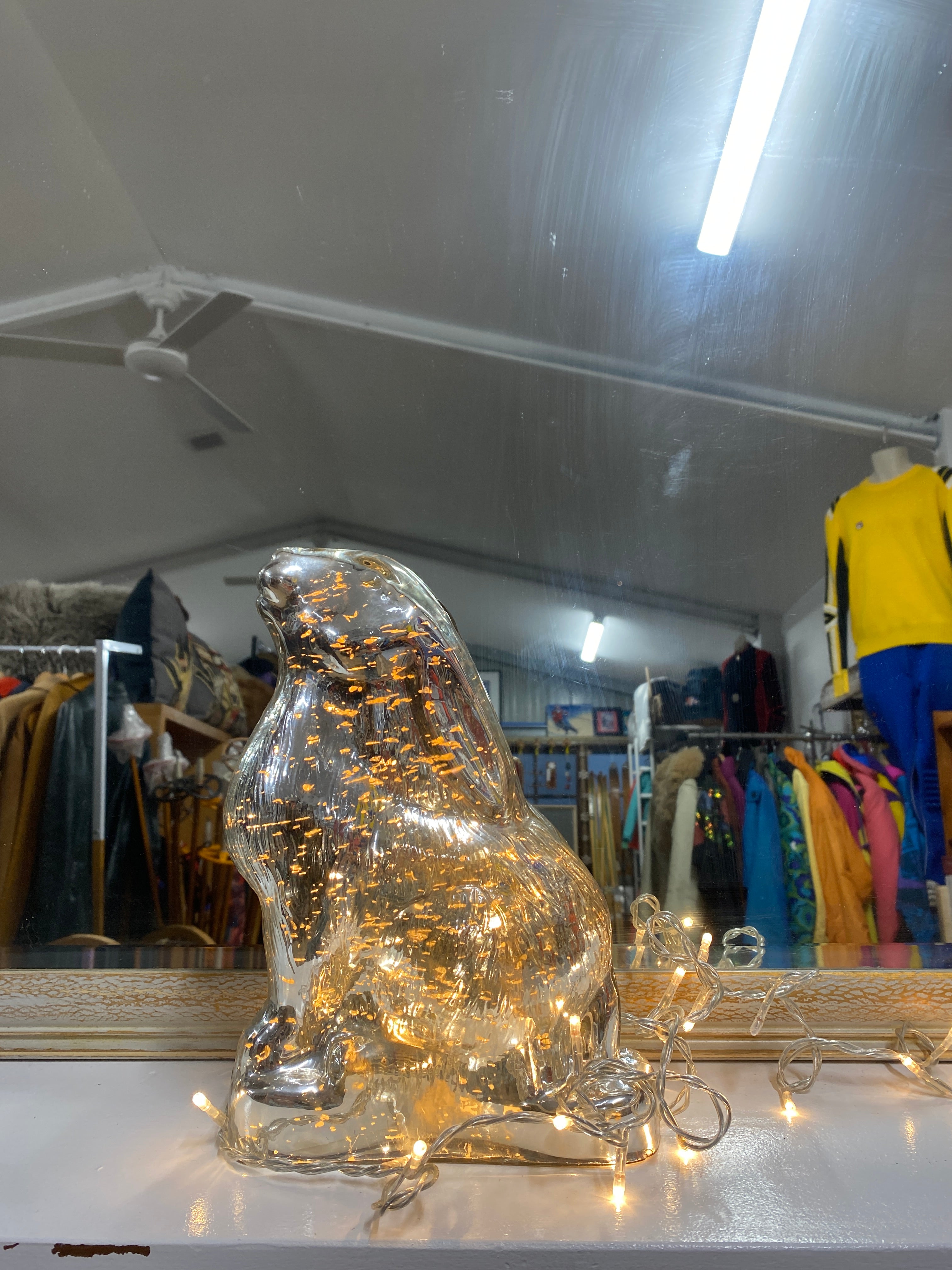 Image shows a silver glass rabbit with fairy lights switched on and displayed externally. The rabbit is sitting on its haunches with it's nose in the air, against a mirror backdrop reflecting vintage snow clothing & sitting on a white bench. Rabbit viewed from the side.