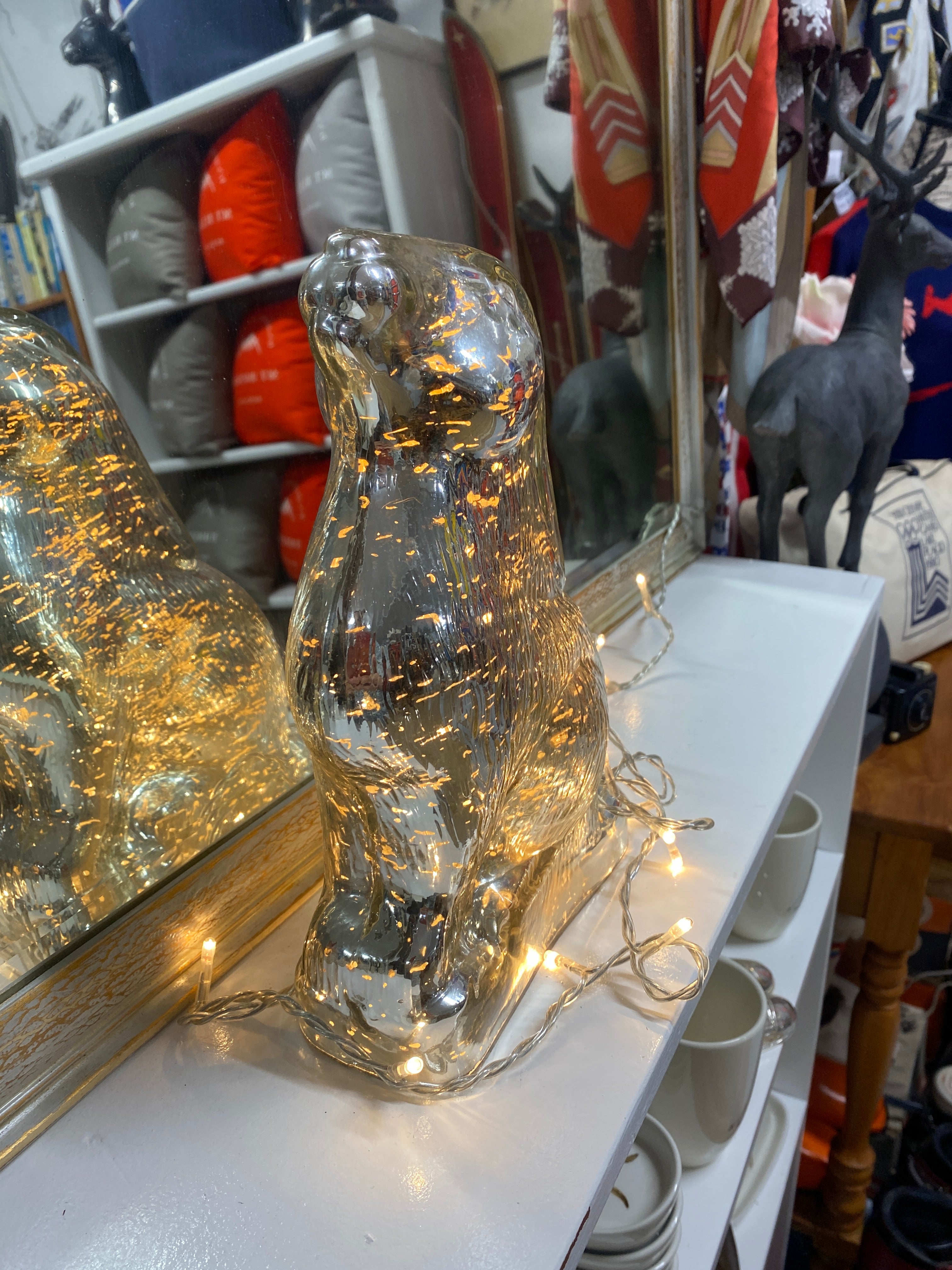 Image shows a silver glass rabbit with fairy lights switched on and displayed externally. The rabbit is sitting on its haunches with it's nose in the air, against a mirror backdrop reflecting vintage snow clothing, cushions and ornaments & sitting on a white bench. Rabbit viewed from the front.
