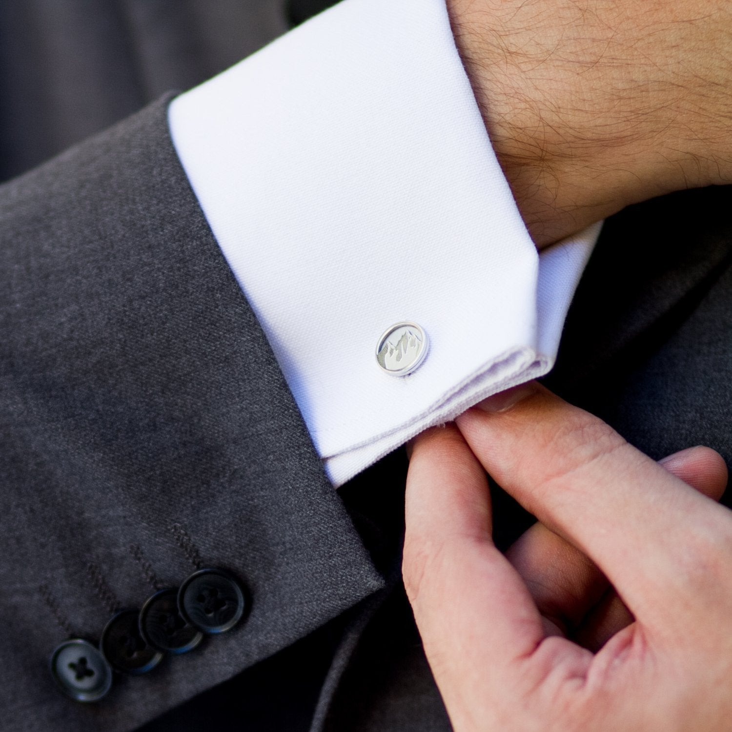 Image shows a person in a grey suit with a white shirt adjusting their shirt cuffs and showing a pair of stainless steel round cufflinks with mountains engraved into their surface.