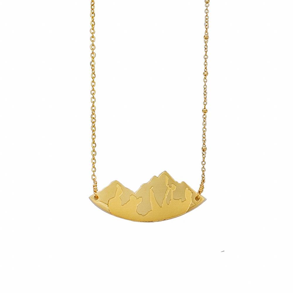 Image shows a gold necklace with pendant detailing mountain peaks and engraved on the pendant surface, against a white backdrop.