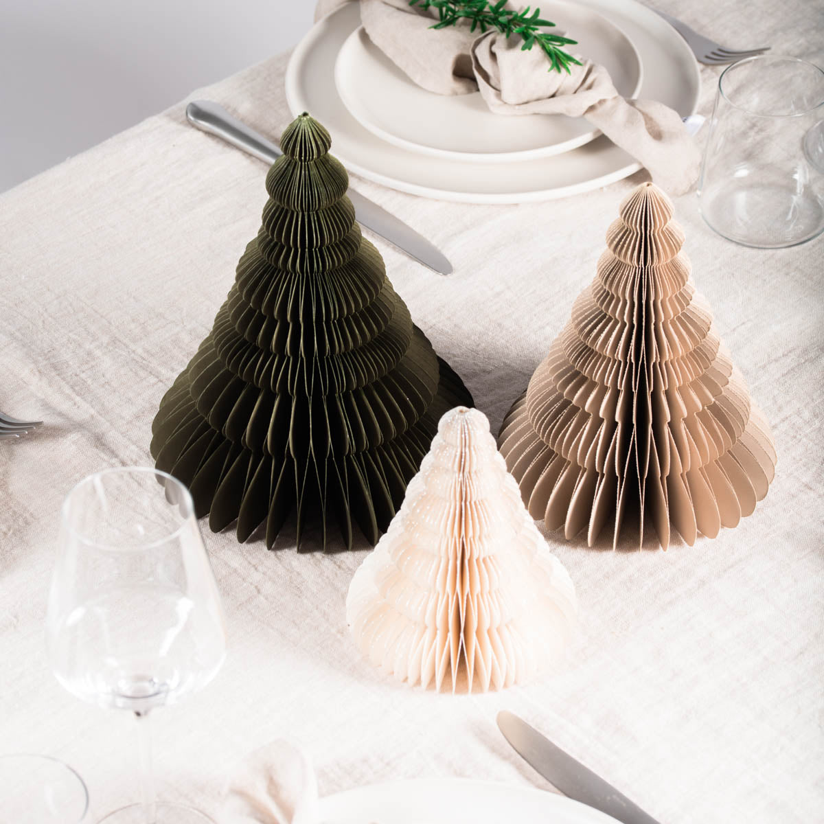 1 Off-White, 1 Olive Green and 1 Linen coloured Paper Standing Christmas Trees, in front of a white painted wall and resting on a table laid with white linen with glassware and rosemary sprigs viewed from above