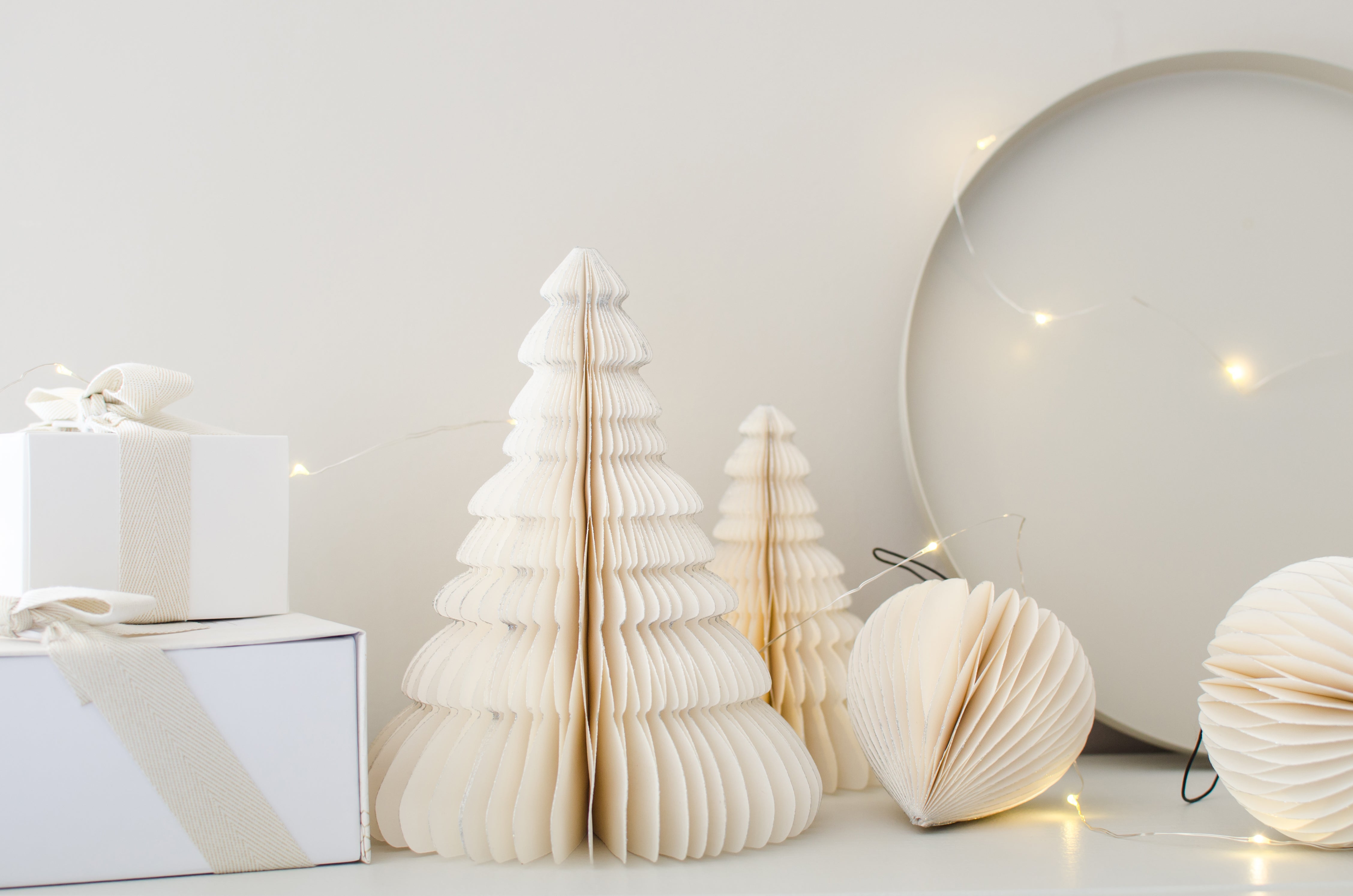 Selection of 2 Off-white Paper Standing Trees, & two hanging paper ornaments in a bauble shape, resting against a white painted wall and table with white gifts boxes and fairy lights
