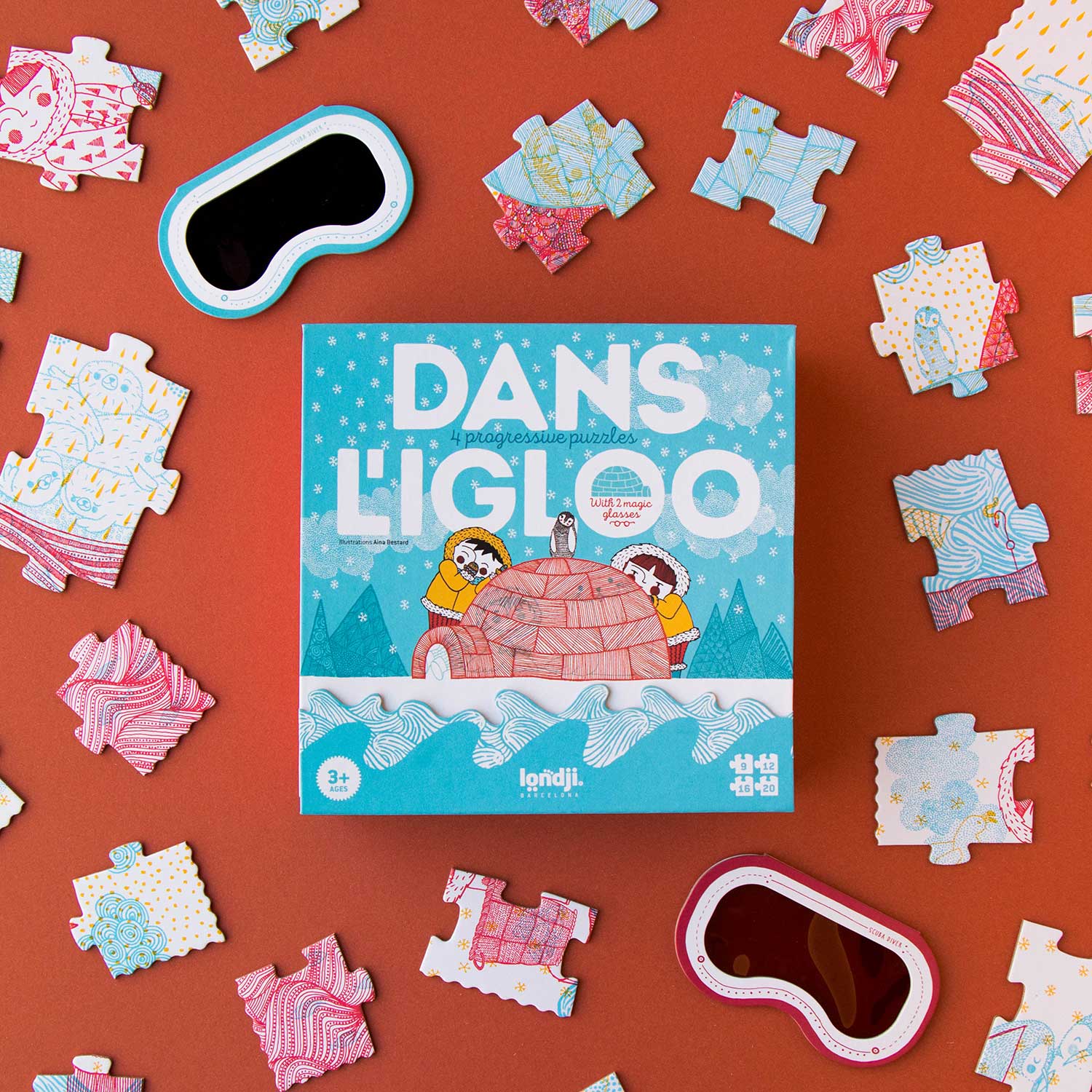 Image shows the front of a square puzzle box named dan's L'igloo, shown from above. Scatters around are puzzle pieces and two 3D viewing screens shaped as goggles. Shown against a red backdrop.