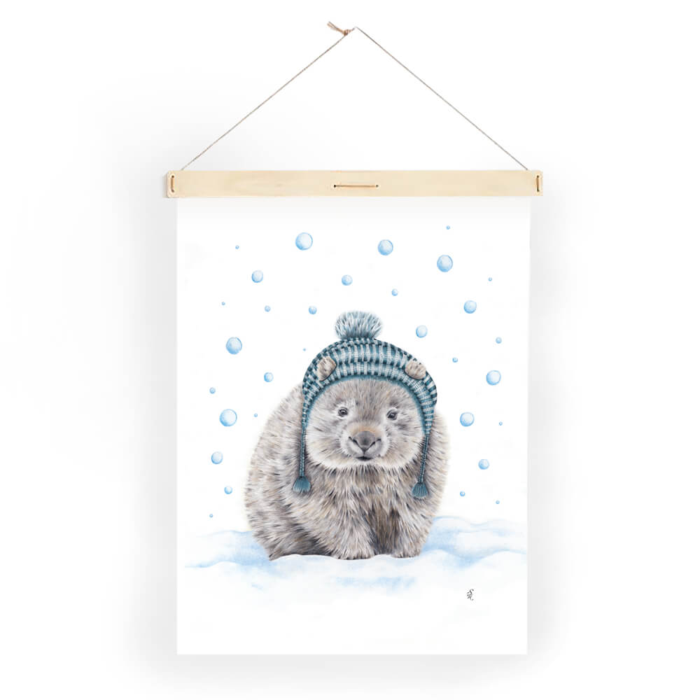 Image shows a tea towel illustrated with a wombat wearing a blue tasseled beanie, standing in blue tinged snow while the snow falls. The tea towel is hanging on a wall using a pale wooden print hanging baton against a white backdrop.