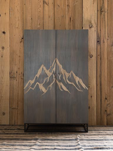 Wooden Cabinet engraved with a mountain & brass ski shaped handles with square metal base & legs on a wood panel background with rug underneath