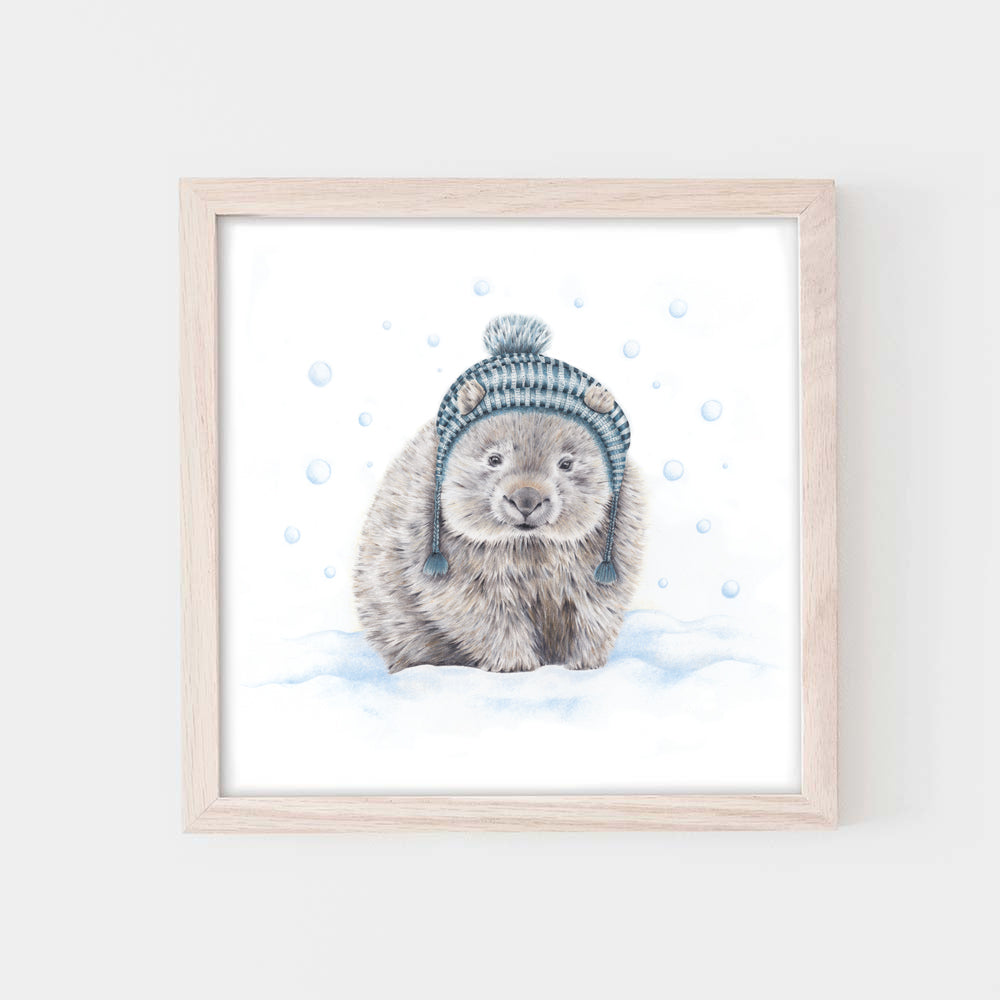 Image shows a illustration of a wombat wearing a blue tasseled beanie, standing in blue tinged snow while the snow falls, framed in pale wood. The frame is hanging on a light grey wall.