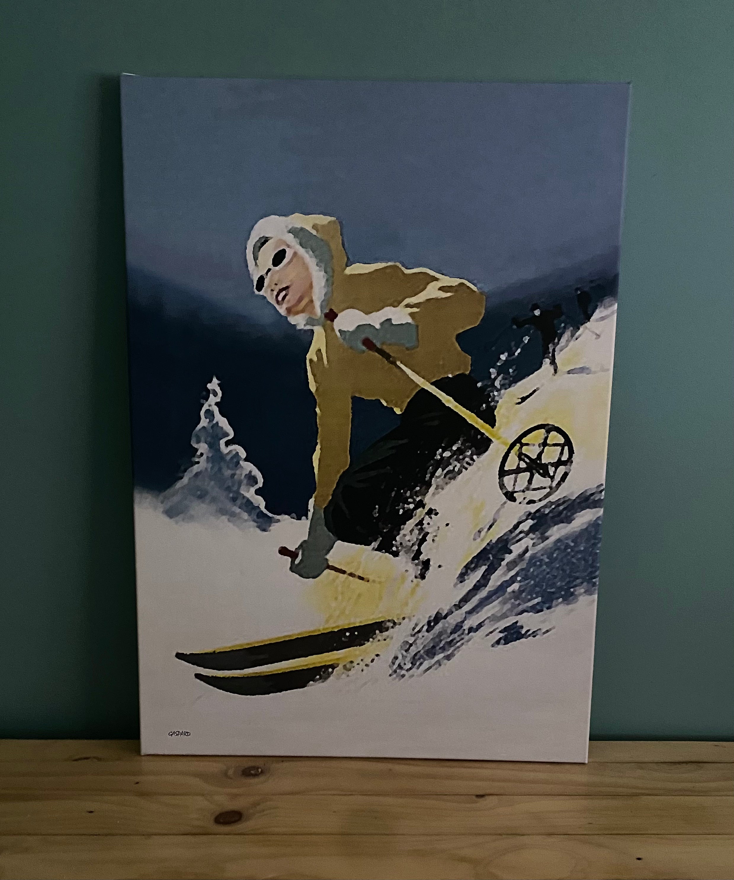 Canvas depicting a woman in a yellow jacket with a white fur at the edge of the hood and blue pants skiing downhill, leaning to the right into the hill with a tree and the shadow of the mountains in the background below a blue sky. Resting against a green wall on a timber table.