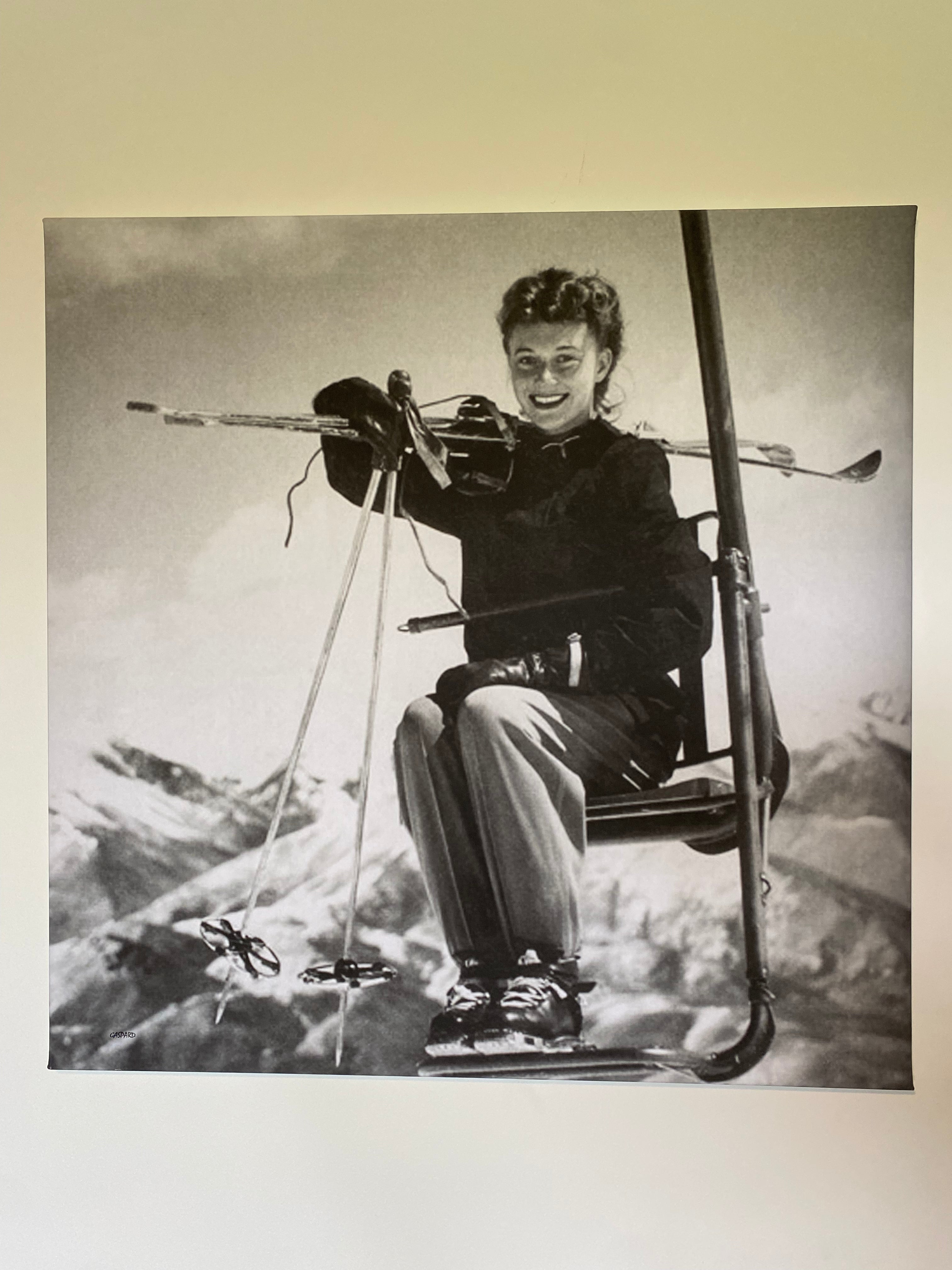 A black and white vintage photo depicting a smiling woman wearing a dark coloured ski jacket, light coloured ski pants and dark leather lace up ski boots,  sitting on a single seat chairlift in foreground with wooden skis and poles on her shoulders looking directly at the camera, with snow covered mountains in the background. Hanging on a white wall