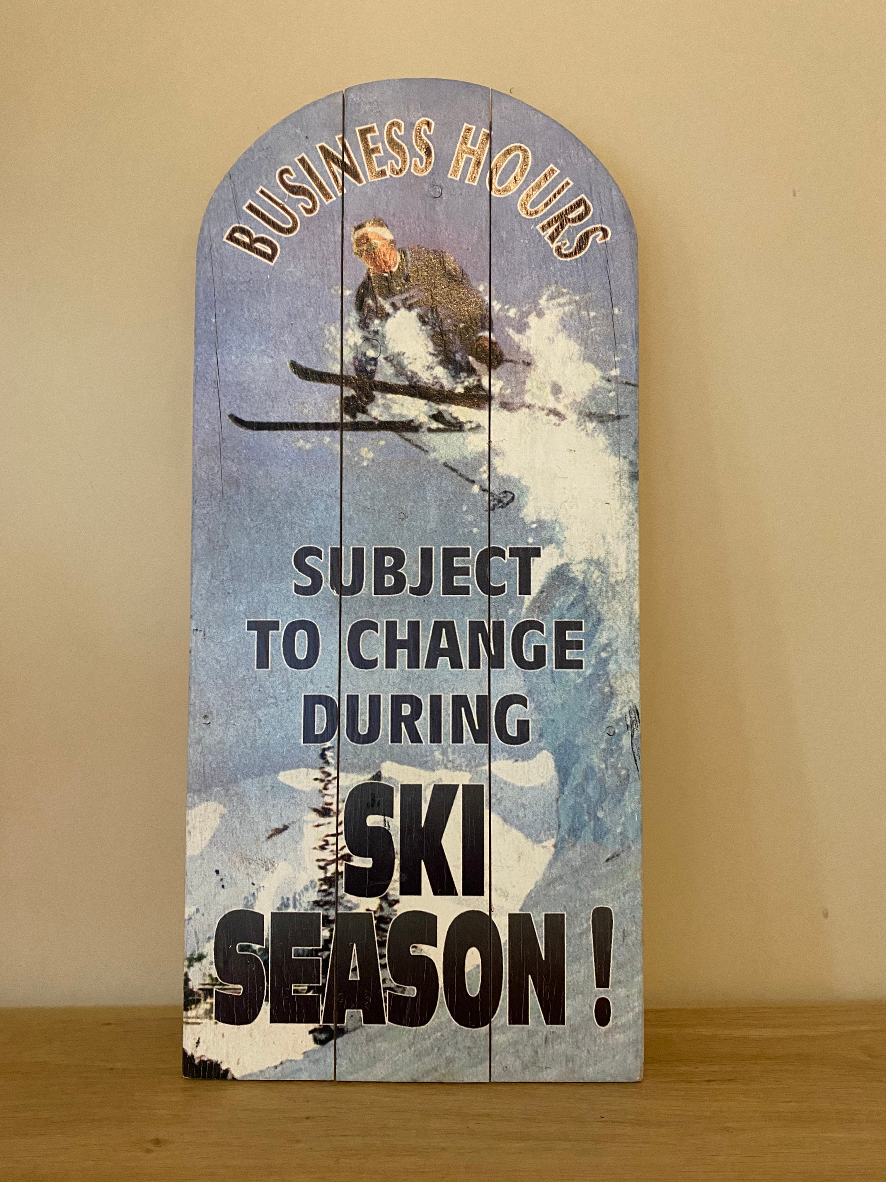 Wooden sign using a vintage photo as the background, showing a skier launching from a snow ledge and soaring into the air in a crouched position. Front view. Resting against a beige wall on a wooden table top