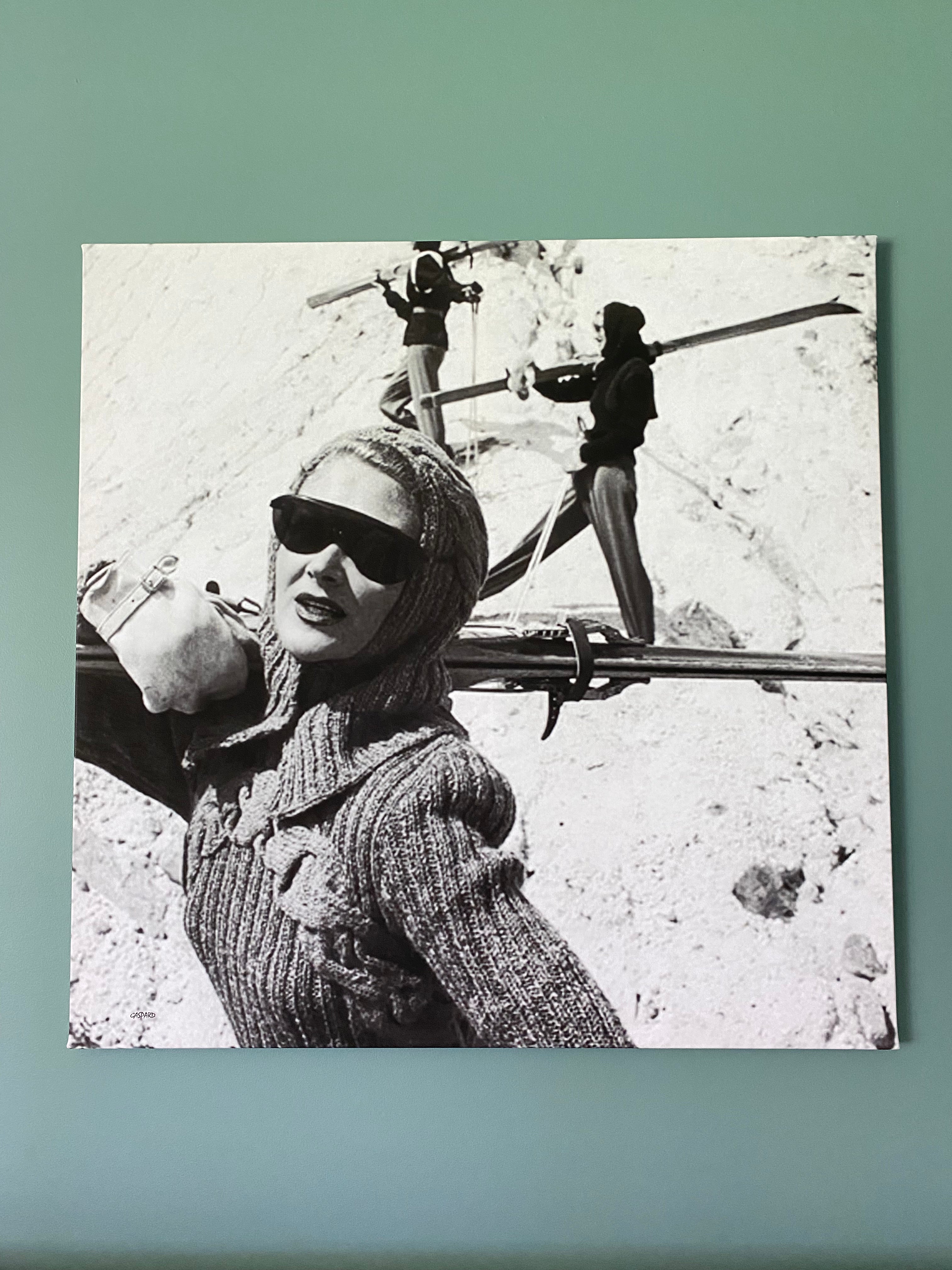 A black and white vintage photo depicting a woman in sunglasses wearing a knitted jumper and a snug hooded cowl in foreground with skis on her shoulders looking directly at the camera, with two women in the background standing with wooden skis on their shoulders on a snowy tracked out slope.  Hanging on a green wall.