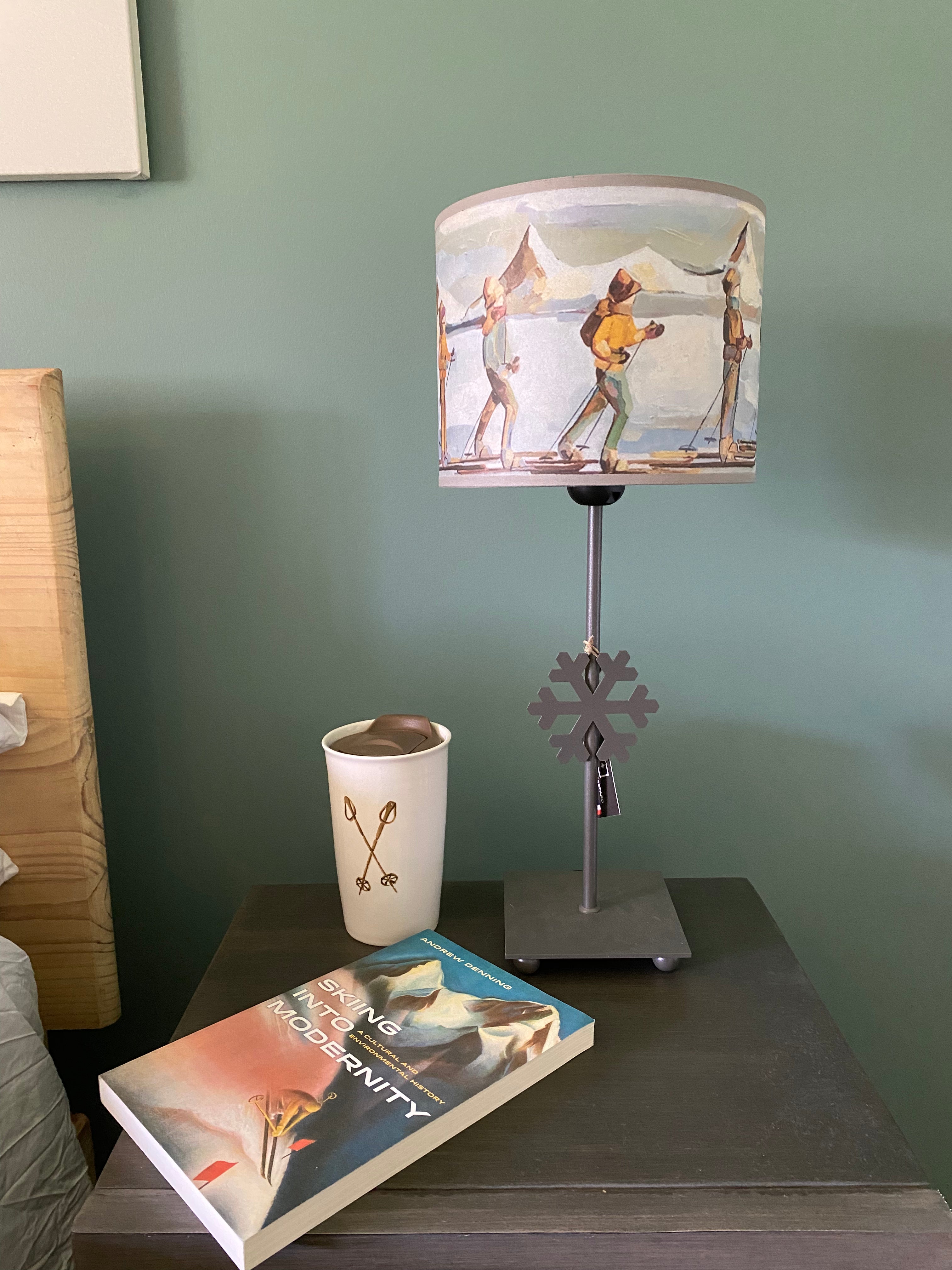 lamp with a square grey solid metal base supporting a rod with ca snowflake mid way up the rod, topped by a colourful canvas lamp shade. The shade depicts people on skis skinning across the snow in a line, one in front of the other, with snow capped mountains in the background. Sitting on a wooden table with a mug with picture of vintage poles, and the book "skiing into Modernity in front of a green wall, beside a bed.
